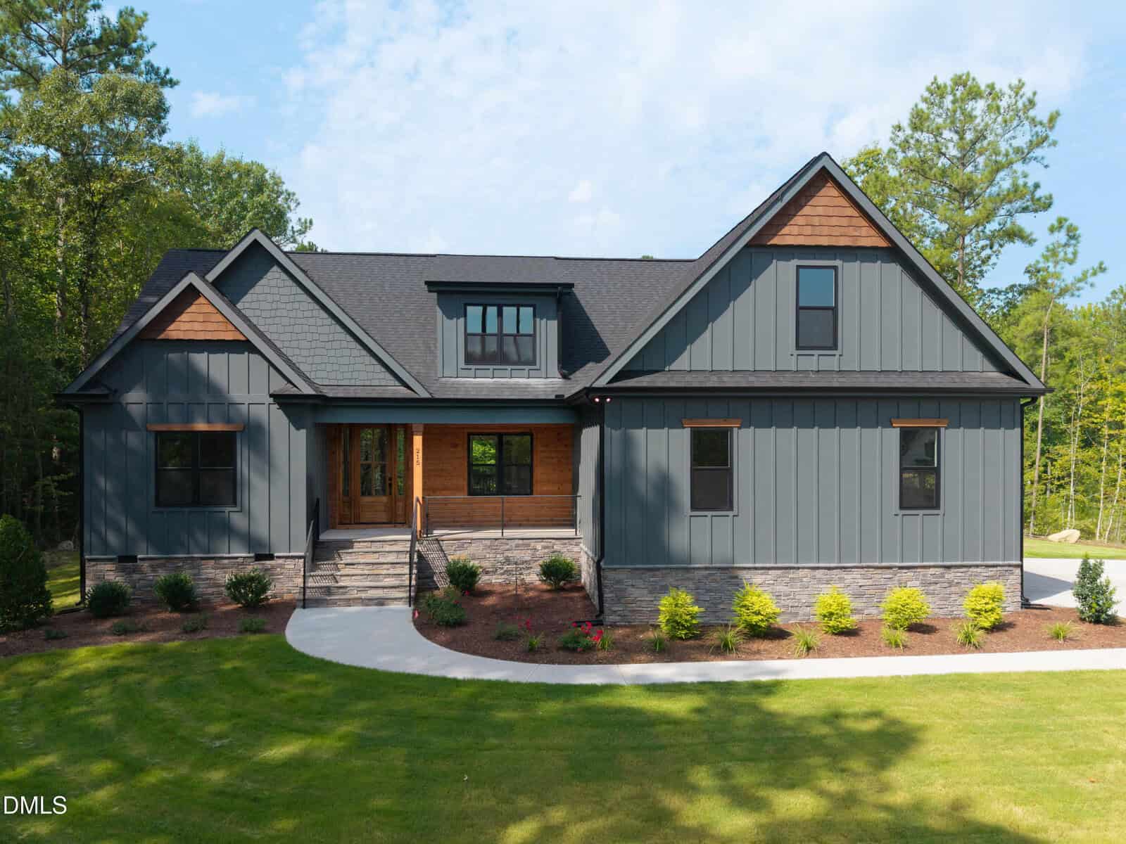 Modern gray and brown house with a gabled roof, stone accents, and wooden front door, surrounded by green lawn, shrubs, and trees on a sunny day—this charming residence at 215 Whistlers Cove in Louisburg perfectly blends style and comfort.