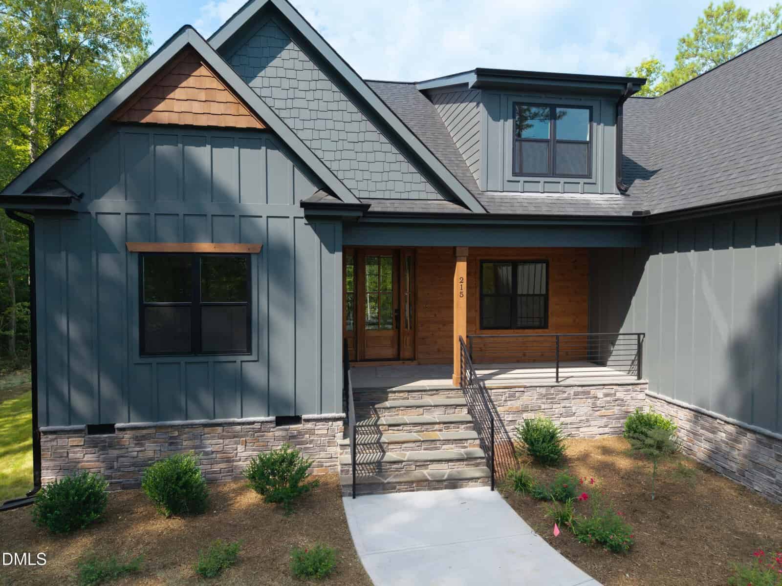 Modern house with dark gray siding, stone accents, wood trim, and covered front porch. Stone steps lead to glass double doors. Small shrubs and mulch line the walkway. Trees are visible in the background.