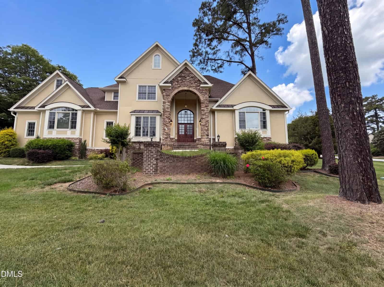 A large beige house with stone accents, arched entryway, and multiple gables, surrounded by green lawn, shrubs, and tall trees under a blue sky with clouds.