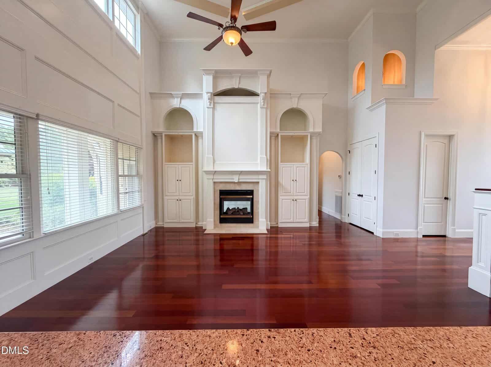 Spacious living room with high ceilings, rich wooden floors, a ceiling fan, built-in white shelves, a fireplace, large windows, and arched alcoves, creating a bright and elegant atmosphere.