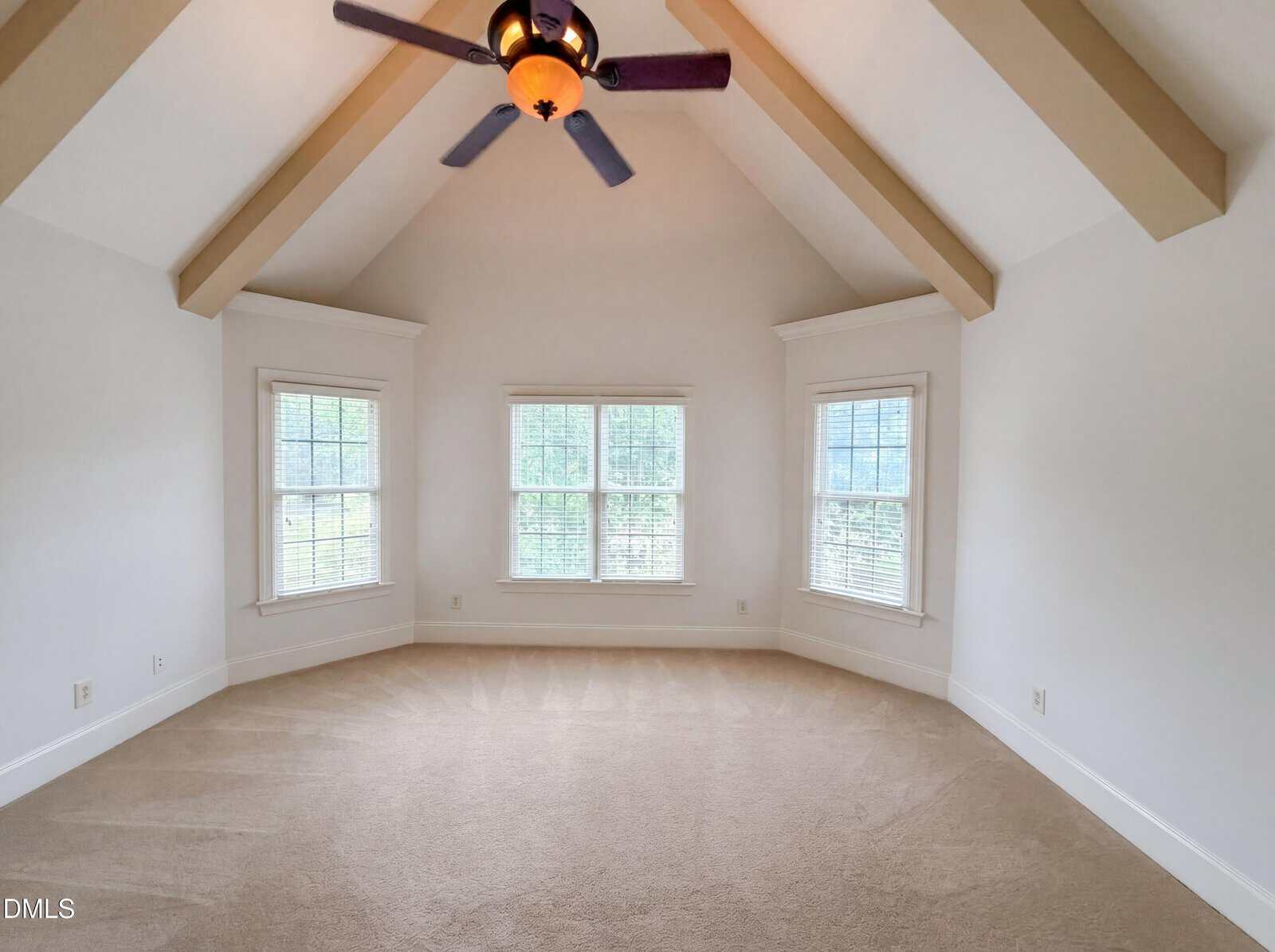 A bright, empty room with beige carpet, white walls, three large windows, and a vaulted ceiling featuring exposed wooden beams and a ceiling fan with a light.