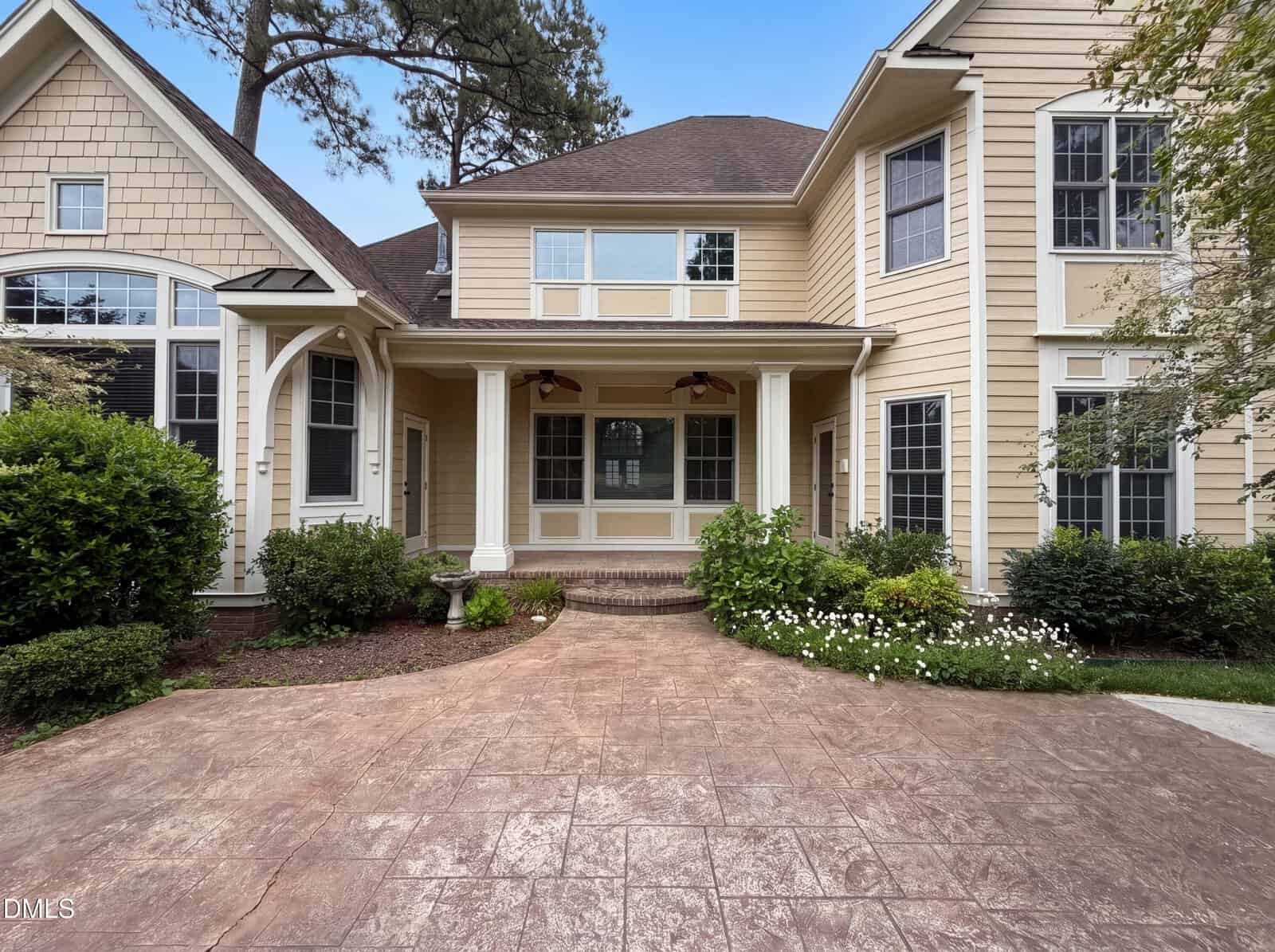 A large, beige two-story house with white trim, brown roof, and multiple tall windows. The entryway has a covered porch with brick steps, surrounded by landscaping and greenery, and a stamped concrete driveway.