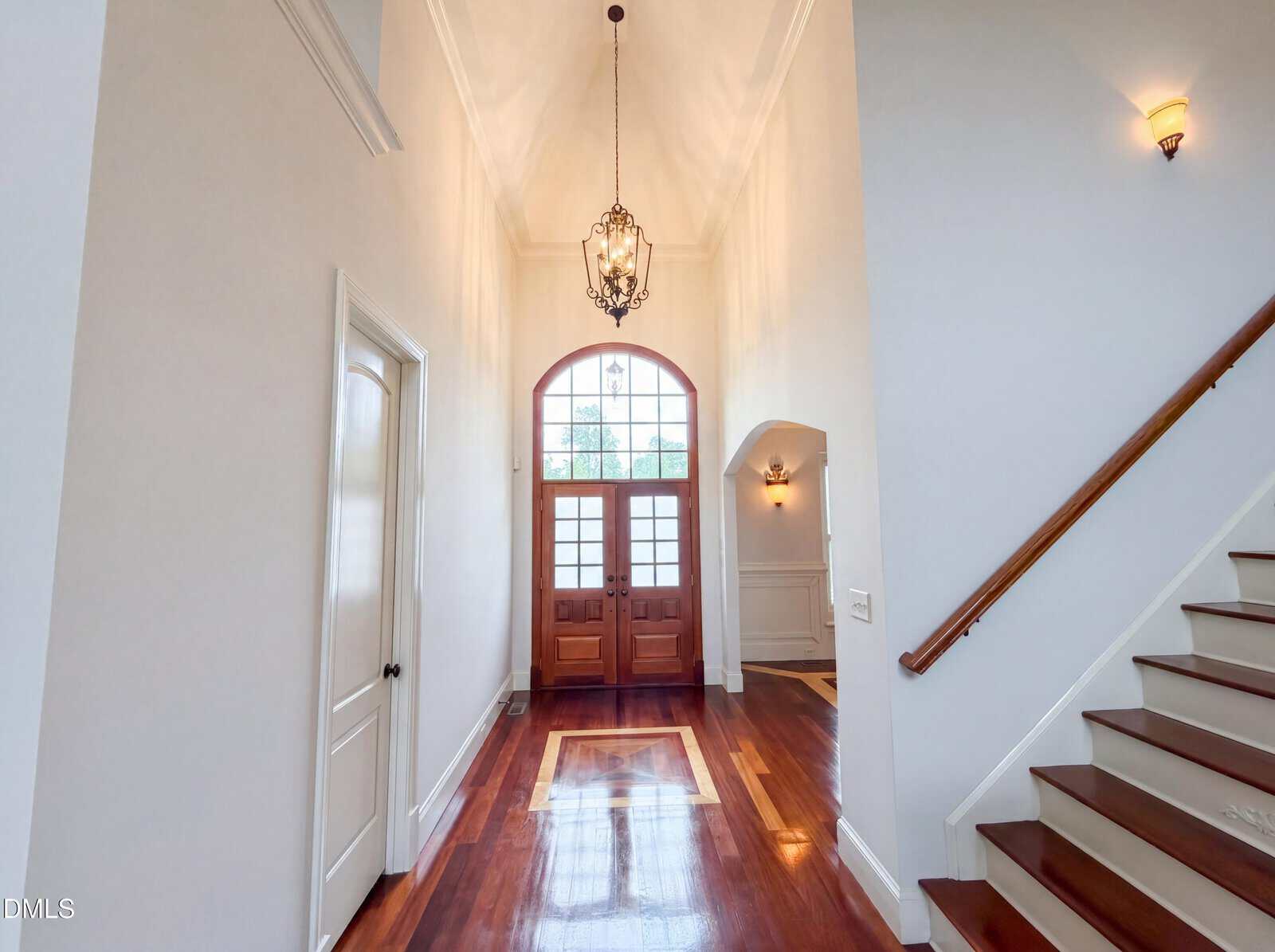 A bright entryway with wooden double doors, large window above, chandelier, white walls, polished wooden floor, and a staircase with a wooden handrail on the right.