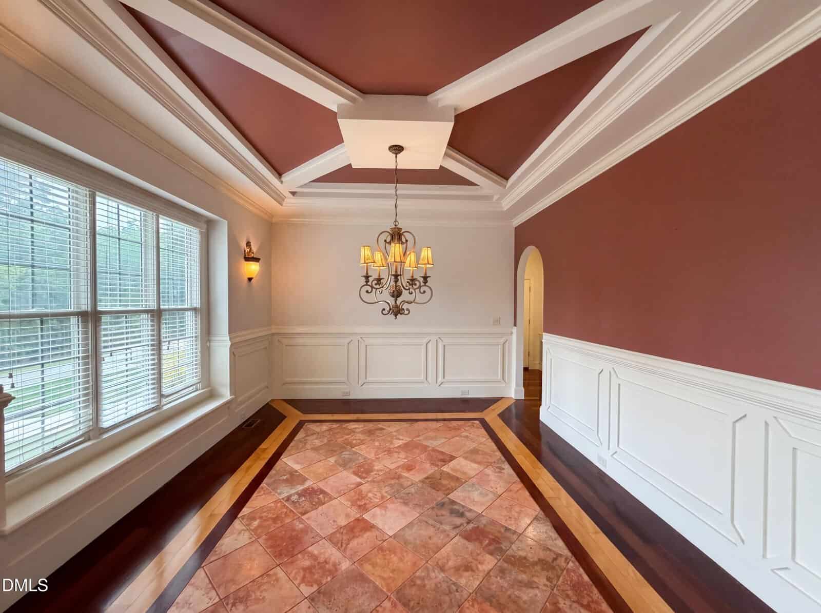 Elegant dining room with a tray ceiling, chandelier, large window, white wainscoting, reddish walls, and a central tile floor design bordered by polished wood. Natural light fills the room.