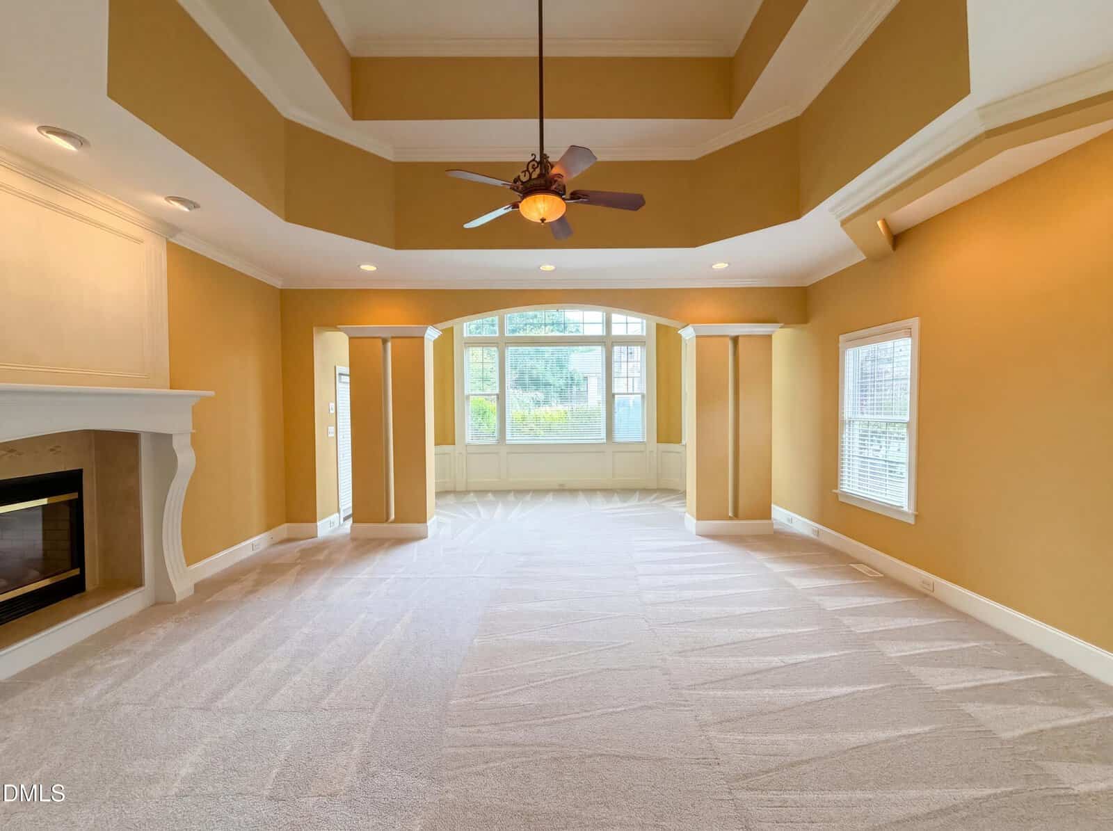 A spacious, carpeted living room with yellow walls, tray ceiling, fireplace, ceiling fan, and large windows overlooking greenery.