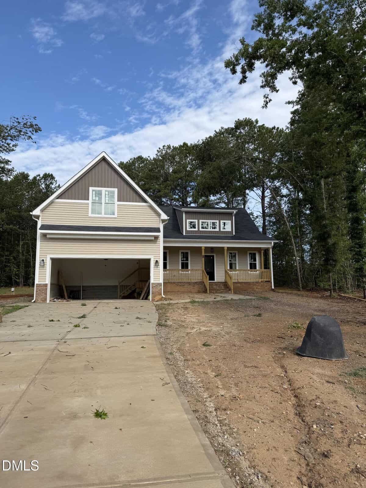 A newly built, two-story house with a covered front porch, double garage, and unfinished yard surrounded by trees, under a partly cloudy sky.