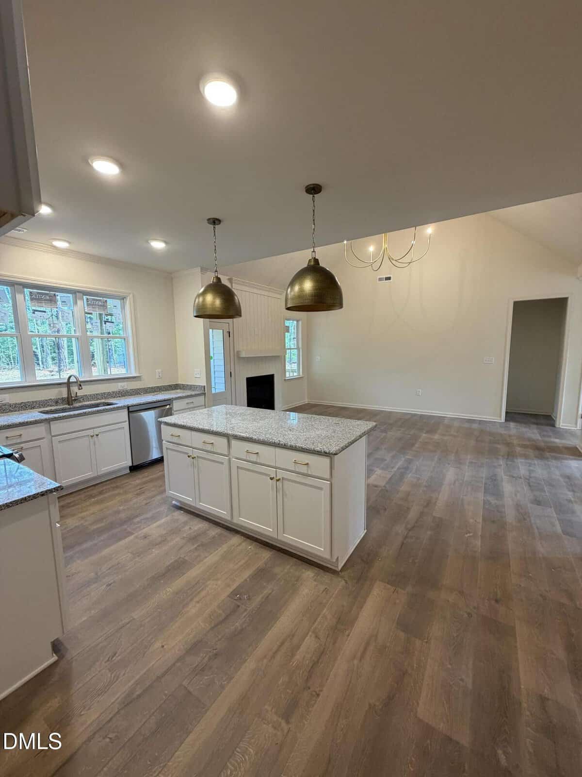 Modern open kitchen with white cabinets, granite countertops, island, pendant lights, and wood flooring, leading to a spacious living area with fireplace and large windows.