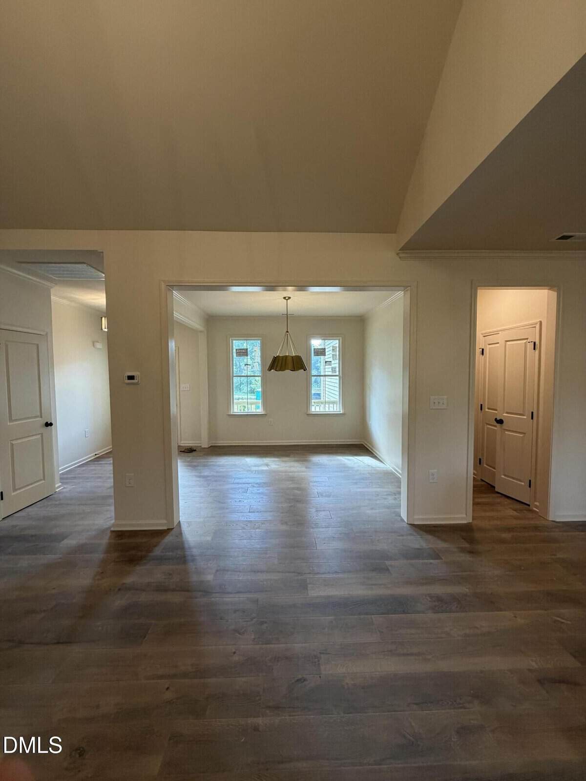View of an empty room with light wood floors, cream walls, a dining area with windows, a modern hanging light fixture, and doorways leading to adjacent rooms. Natural light brightens the space.