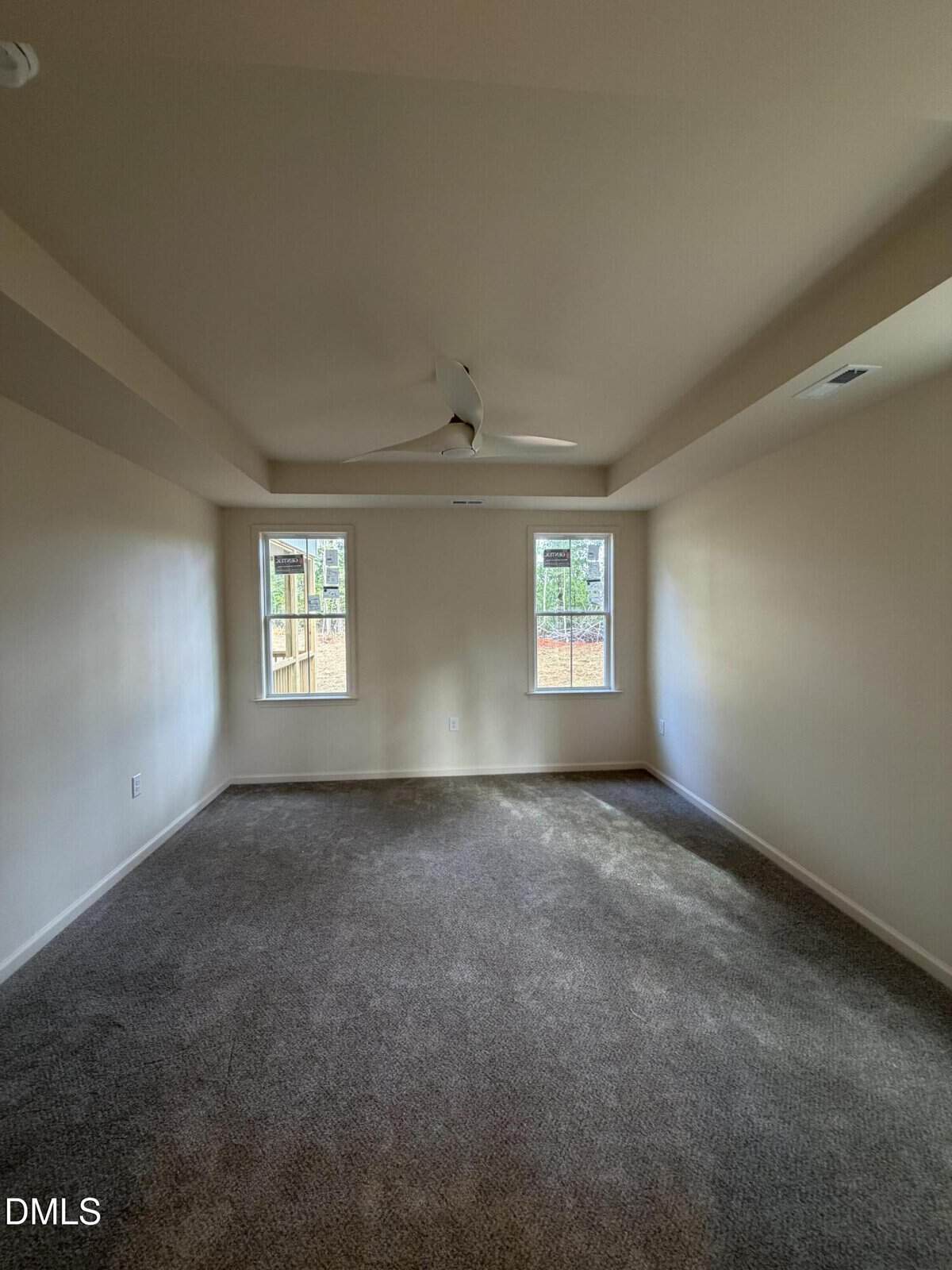 Empty room with beige walls, two tall windows, gray carpet, a white ceiling fan, and sunlight coming through the windows, creating shadows on the floor.