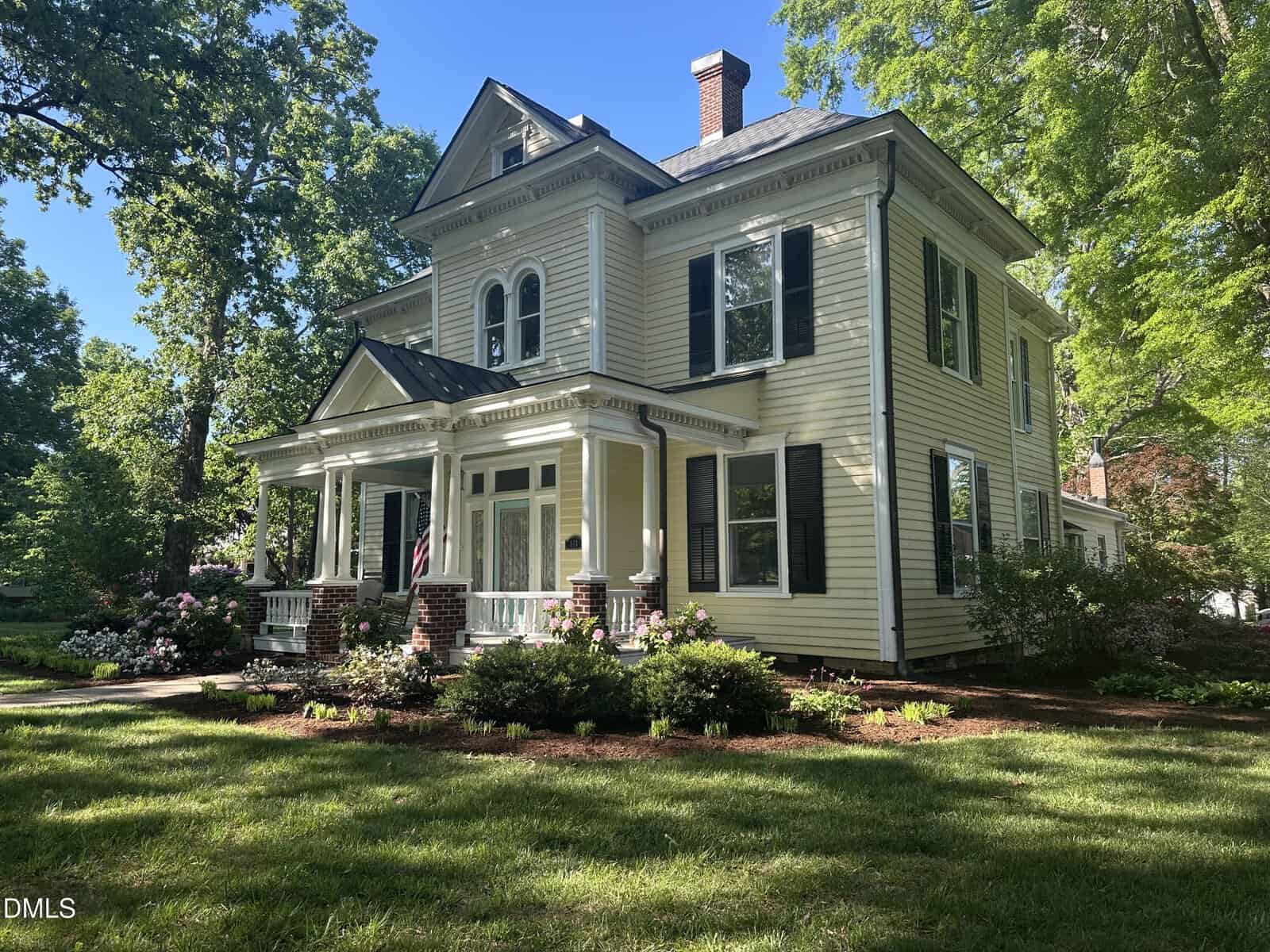 A two-story yellow Victorian-style house with white trim, black shutters, and a covered front porch, surrounded by green grass, bushes, flowers, and tall trees on a sunny day.