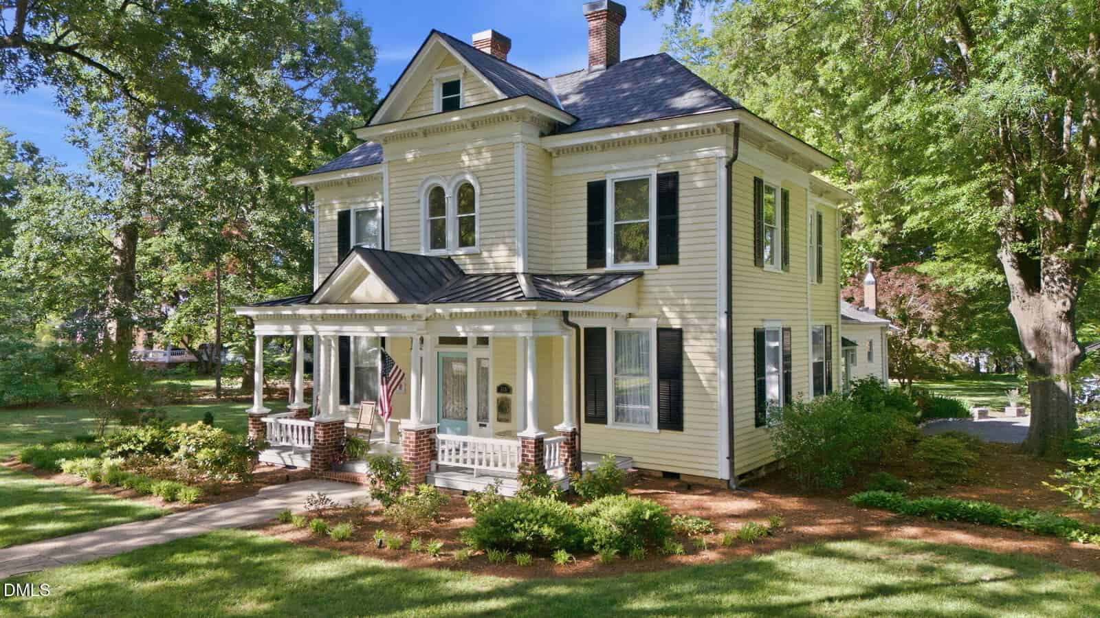 A large, two-story, light yellow Victorian-style house with white trim, a covered front porch, and an American flag, surrounded by green grass, trees, and shrubs on a sunny day.