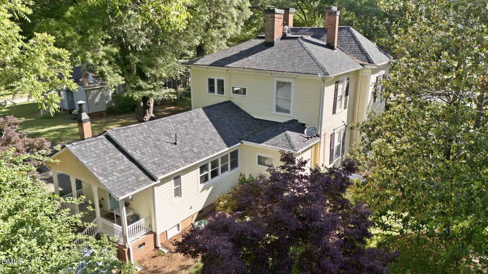 Aerial view of a two-story, light yellow house with a dark gray roof, multiple chimneys, and surrounded by green trees and shrubs. A screened porch is attached to the side of the house.