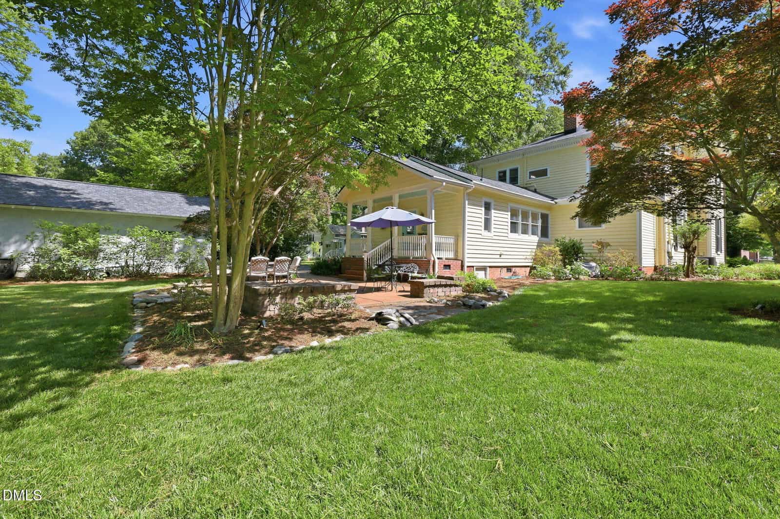 A sunny backyard with green grass, mature trees, and a yellow house. There is a patio with outdoor chairs, a table, and a blue umbrella, surrounded by landscaped garden beds and shaded by trees.