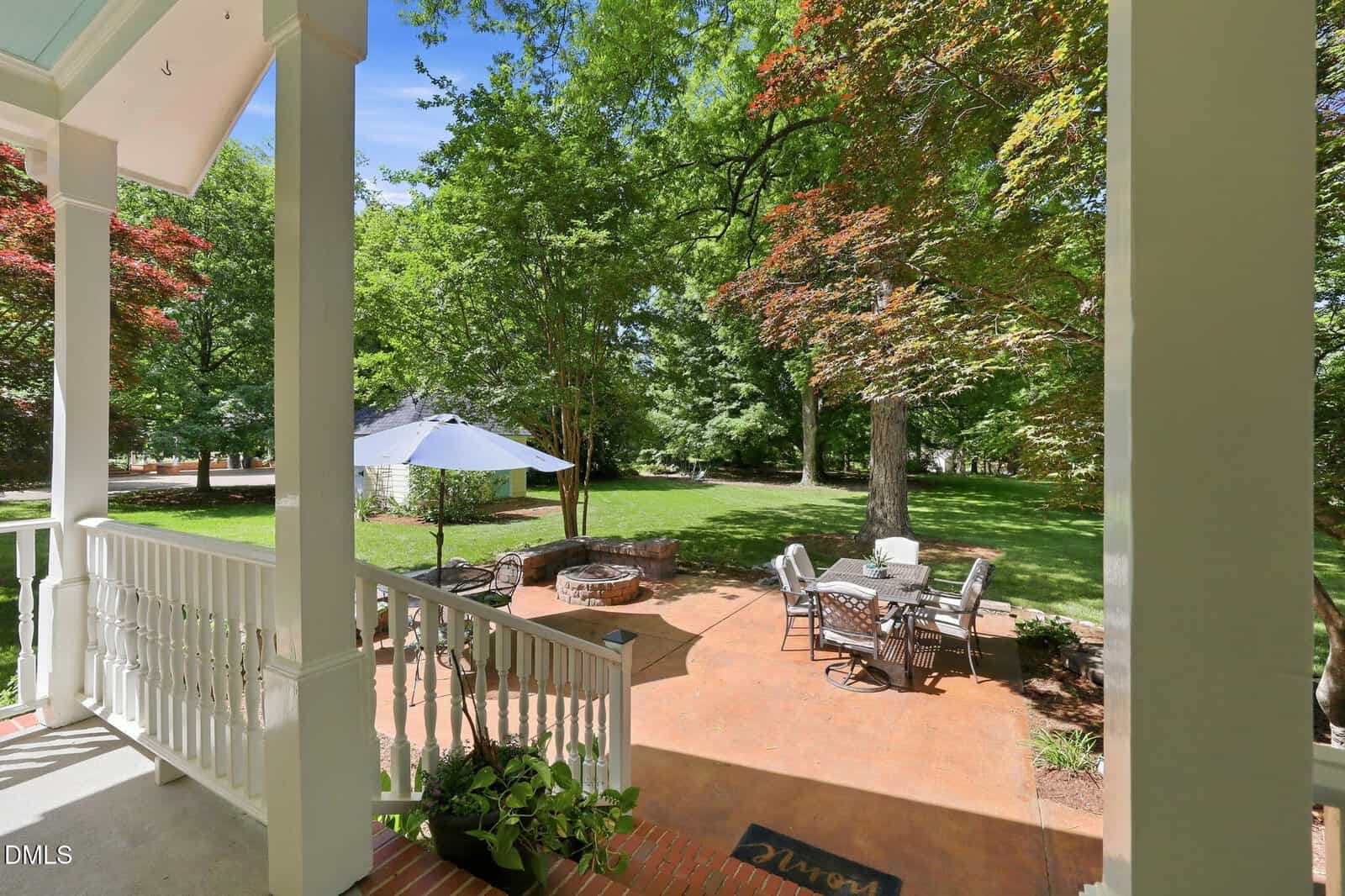 View from a porch overlooking a spacious backyard patio with outdoor dining table, umbrella, fire pit, and surrounded by lush green trees under a clear blue sky.