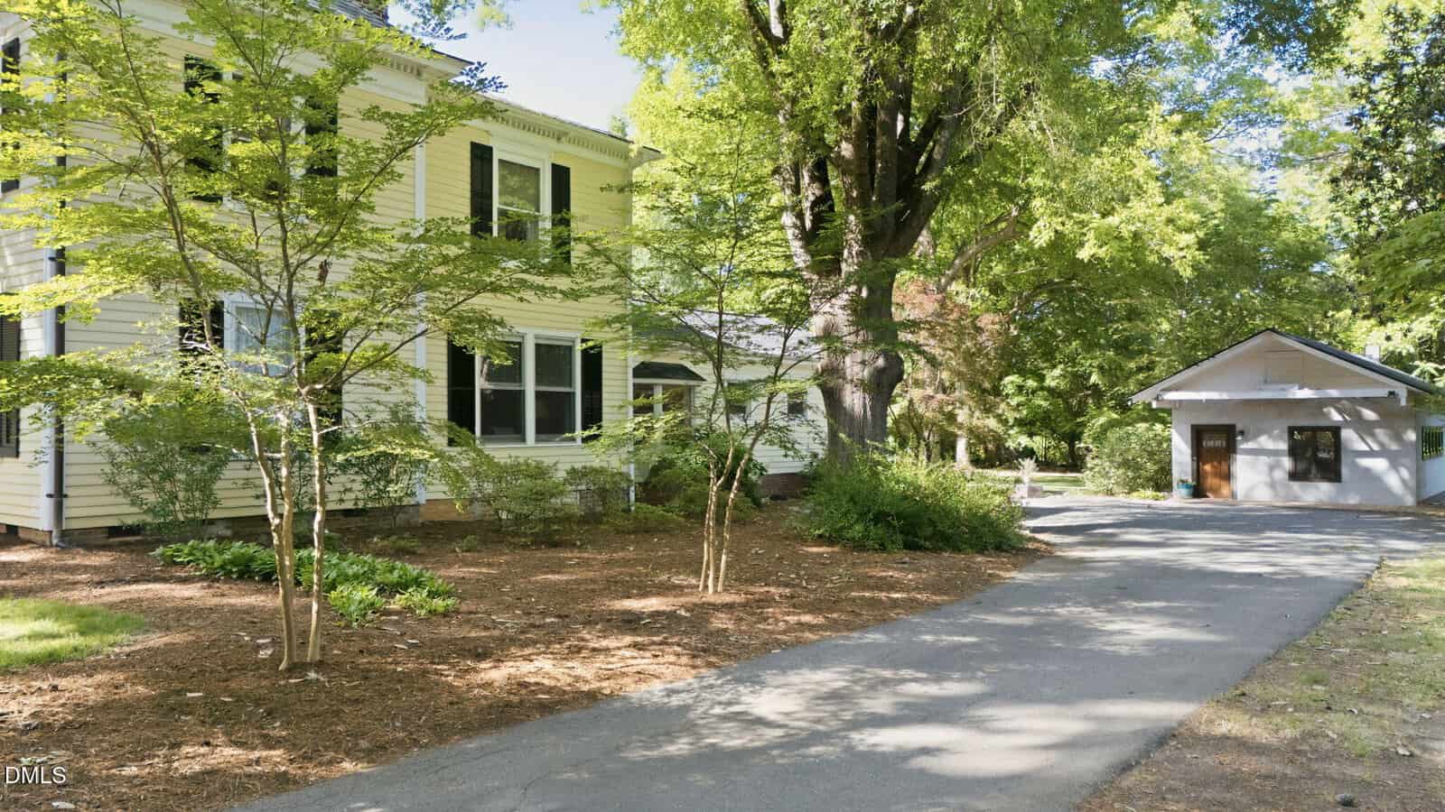 A white two-story house with large windows sits next to a paved driveway leading to a detached one-story garage, surrounded by green trees and landscaped mulch beds.