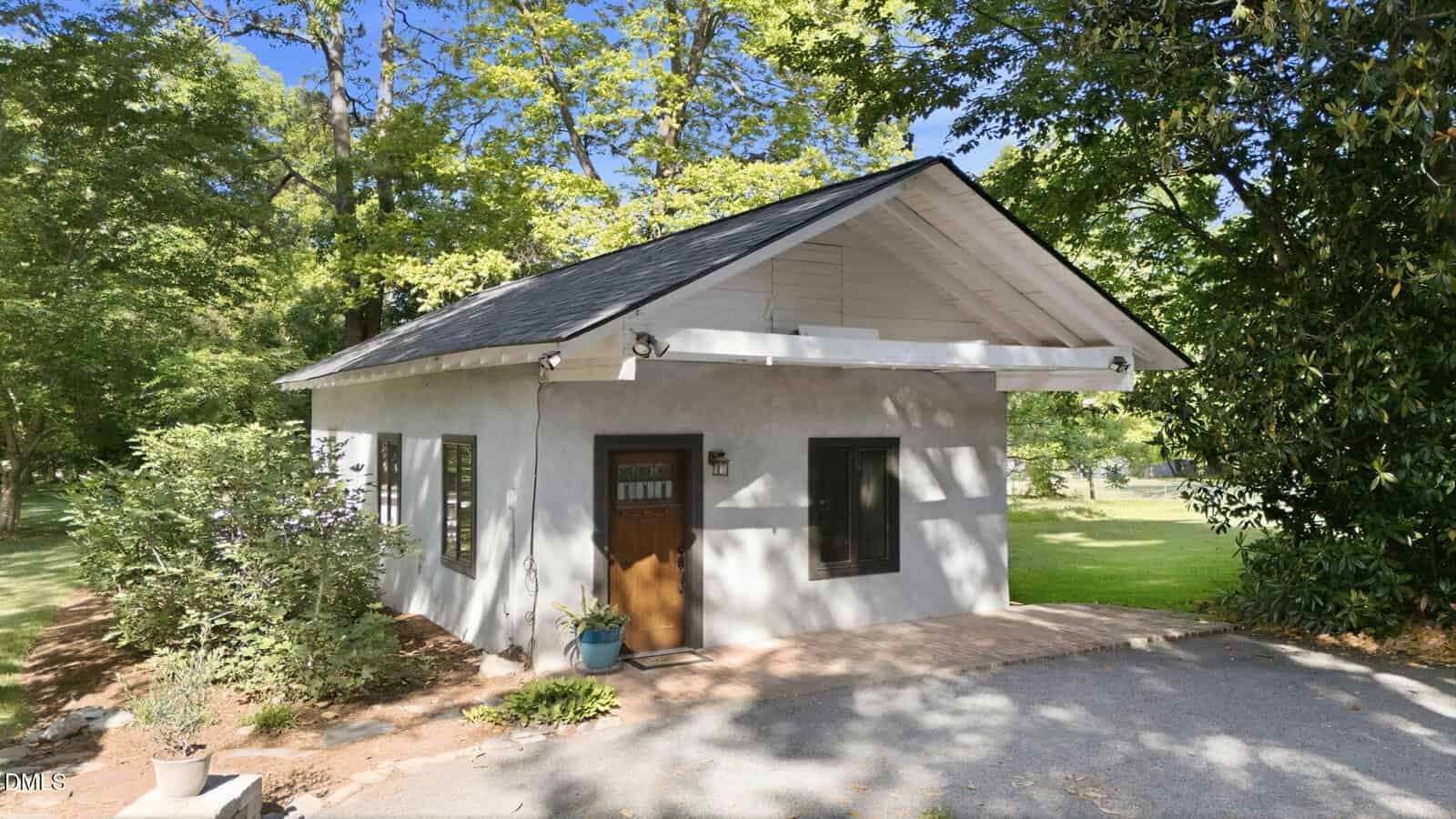Small, light-gray house with a gabled roof, wooden front door, and two black-framed windows, surrounded by green trees and grass on a sunny day. A potted plant sits near the entrance.