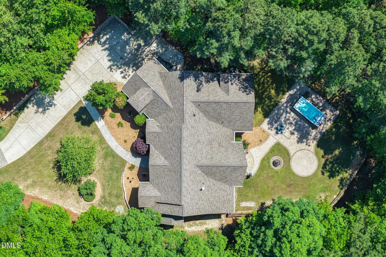 Aerial view of a house surrounded by trees, with a circular driveway on the left and a small pool or hot tub in the backyard. The yard has landscaped paths and garden features.