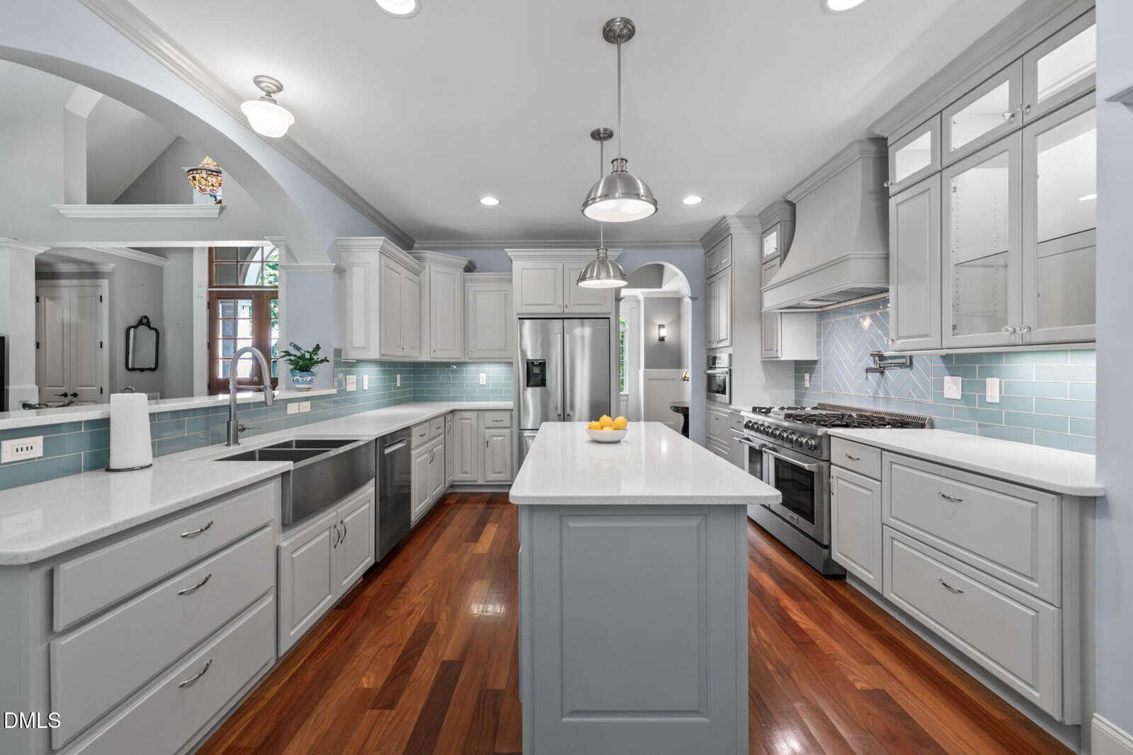 Modern kitchen with white cabinets, light blue backsplash, stainless steel appliances, large island with sink, pendant lights, and wood floors. Arched doorways and open shelving are also visible.