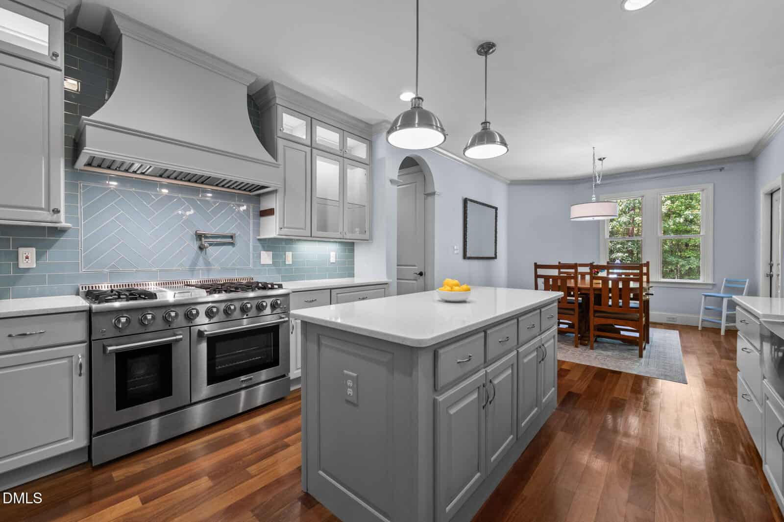 Modern kitchen with stainless steel appliances, blue tile backsplash, gray cabinets, a center island with a bowl of lemons, and an adjoining dining area with a wooden table and chairs near windows.