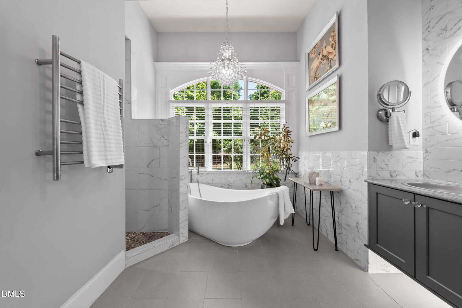 Bright, modern bathroom with a freestanding bathtub by large windows, marble finishes, a walk-in shower, chandelier, vanity with sink, wall art, plants, and a towel rack with white towels.