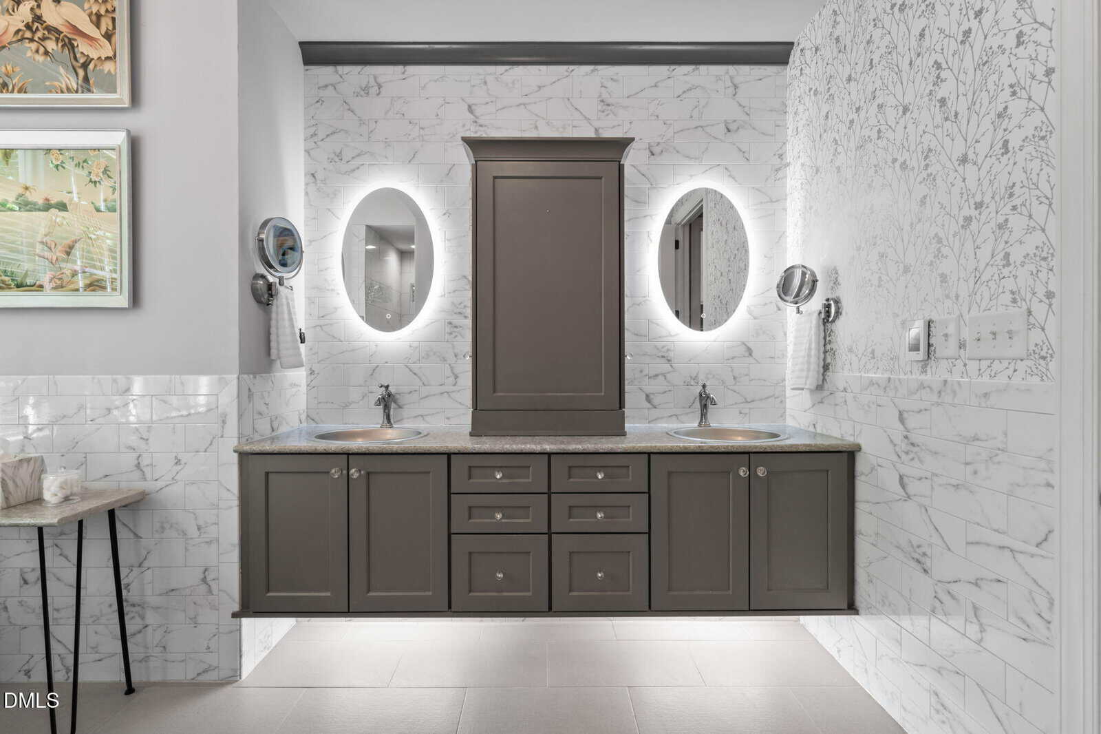 Contemporary bathroom with a double-sink vanity, gray cabinets, marble tile walls, oval backlit mirrors, and a central cabinet. The space features wall-mounted faucets and an elegant, modern design.