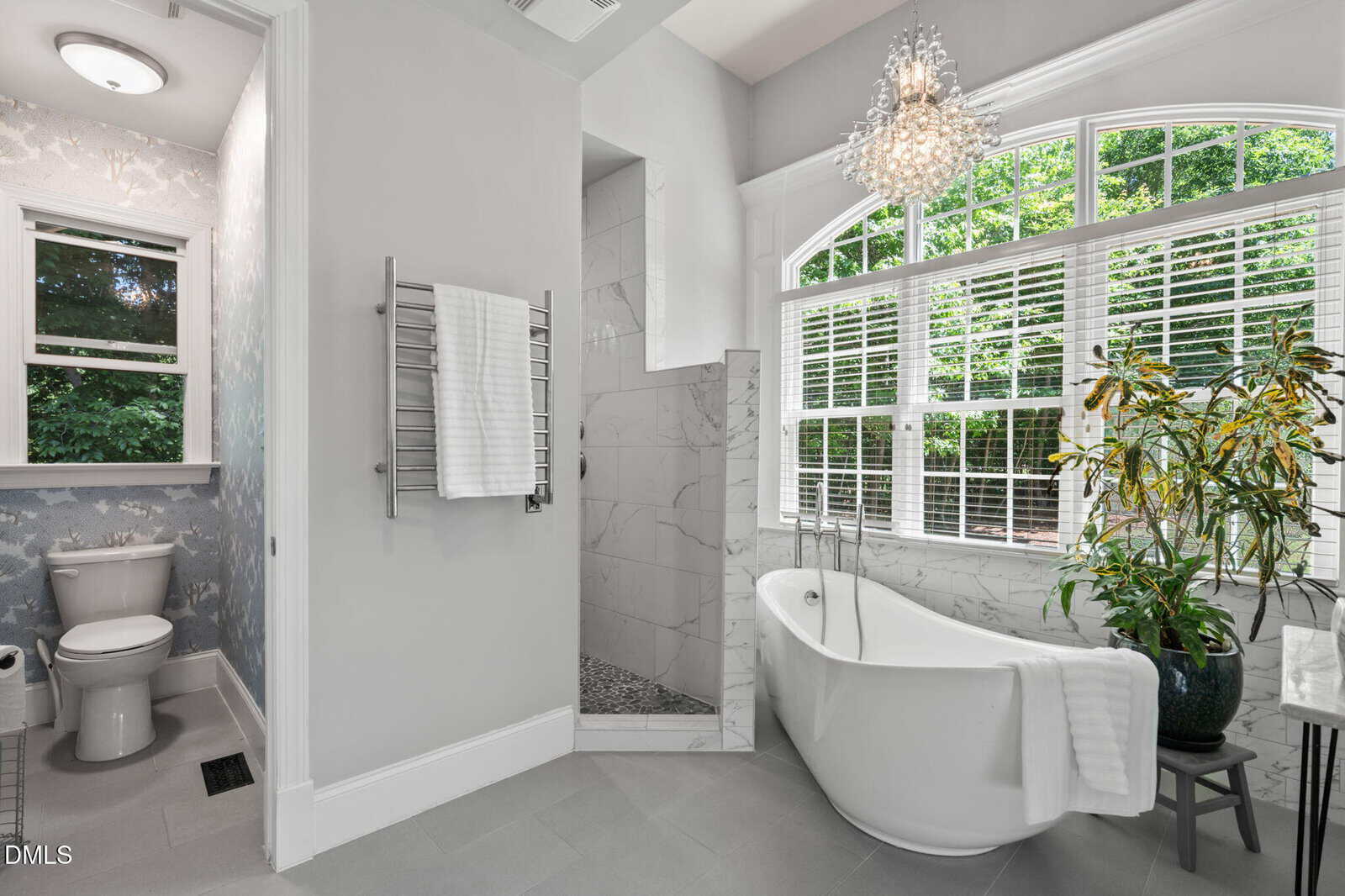 Modern bathroom with freestanding tub by a large window, indoor plant, glass shower, towel warmer, and separate toilet area. Light gray walls, marble surfaces, and a chandelier create a bright, elegant space.