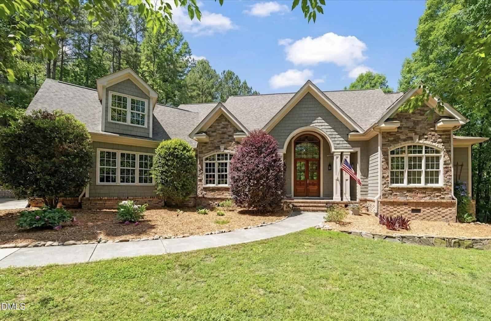 A single-story house with stone and wood siding, gabled roof, large windows, double wooden front doors, manicured shrubs, a small American flag, and a curved sidewalk leading through a landscaped front yard.