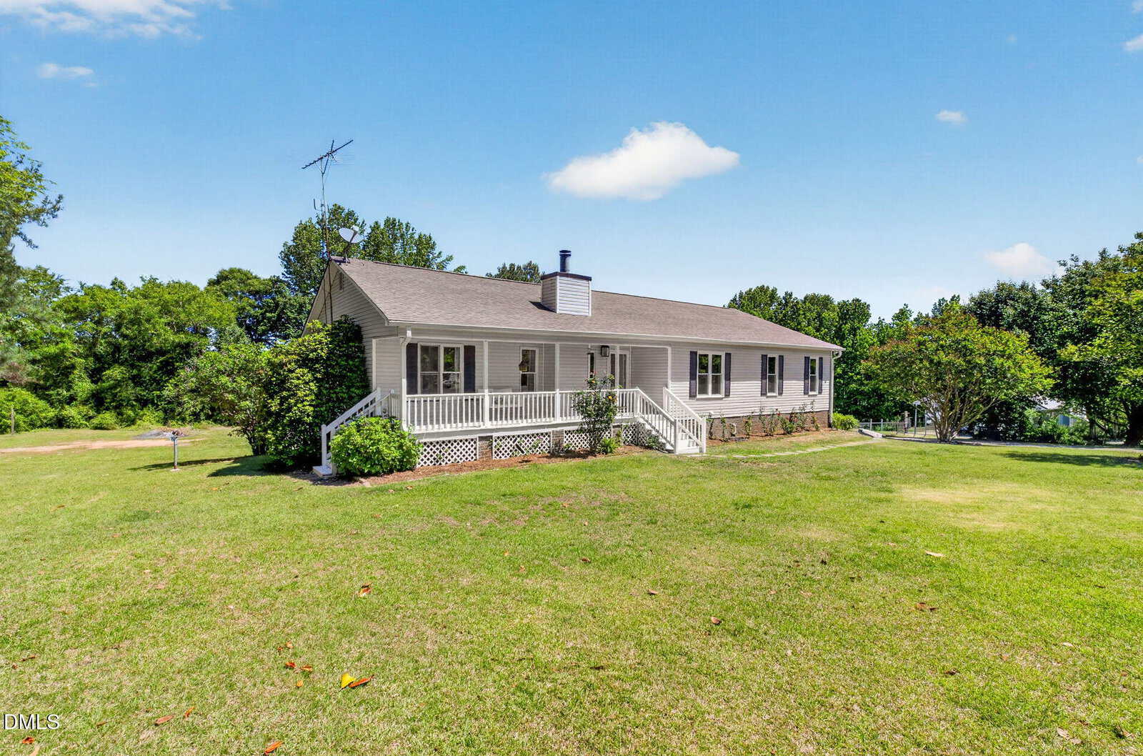 A single-story beige house with a gray roof, white trim, and a front porch sits on a large, grassy lawn surrounded by trees under a clear blue sky.