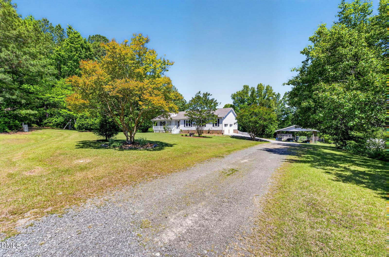 A white house with a porch sits surrounded by trees, with a gravel driveway leading up to it through a sunny, grassy yard. A detached carport or garage is visible to the right of the house.
