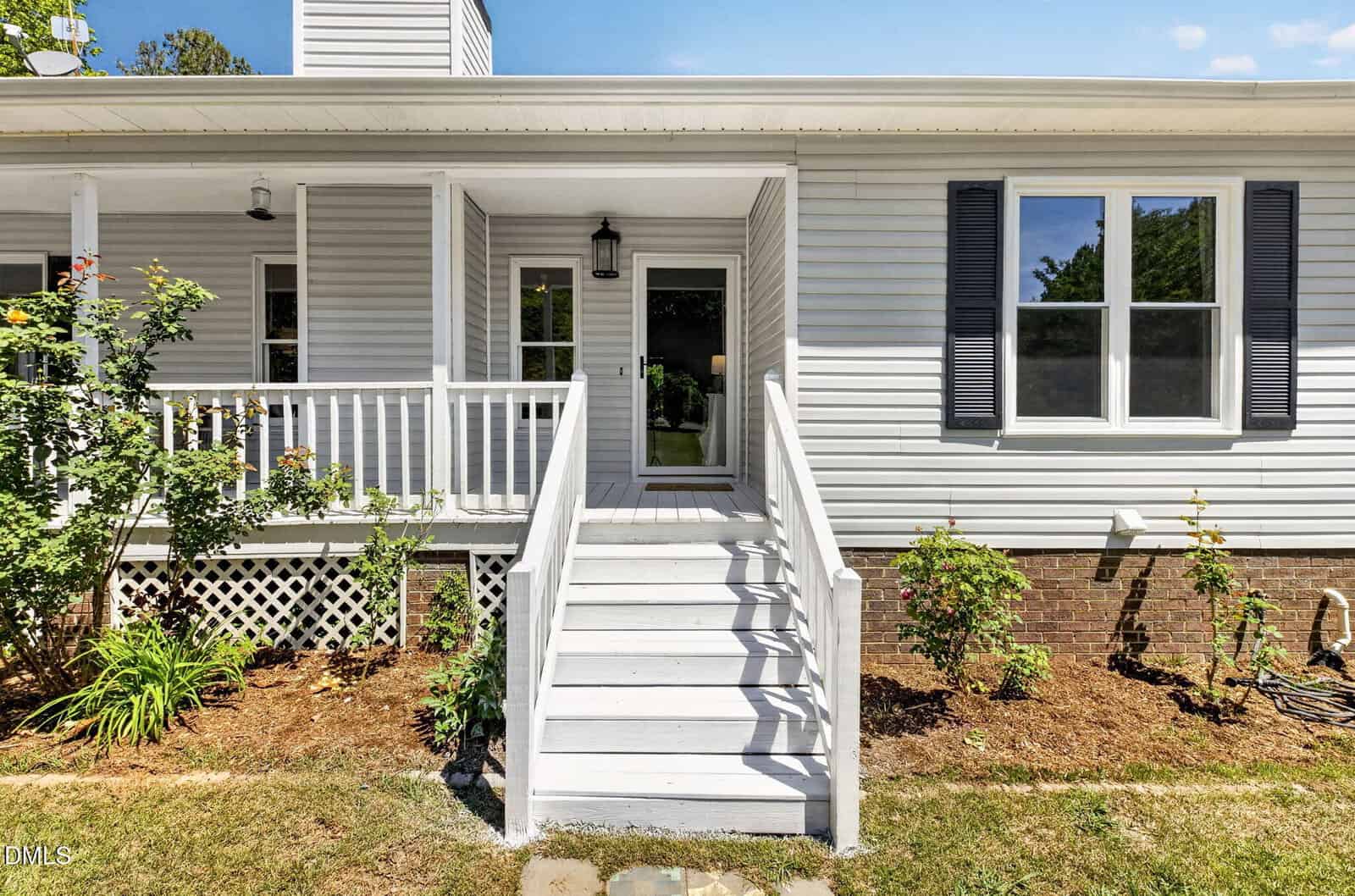 Front view of a white house with a covered porch, central steps leading to a glass door, white railings, dark shutters, brick foundation, and landscaped garden with rose bushes.
