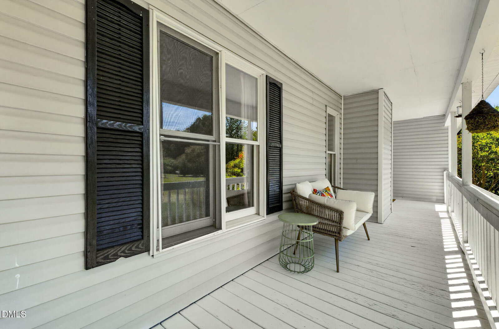 A spacious white porch with a wicker chair, green round side table, and decorative pillow. Black shutters frame the window, and sunlight brightens the space, showing greenery in the background.