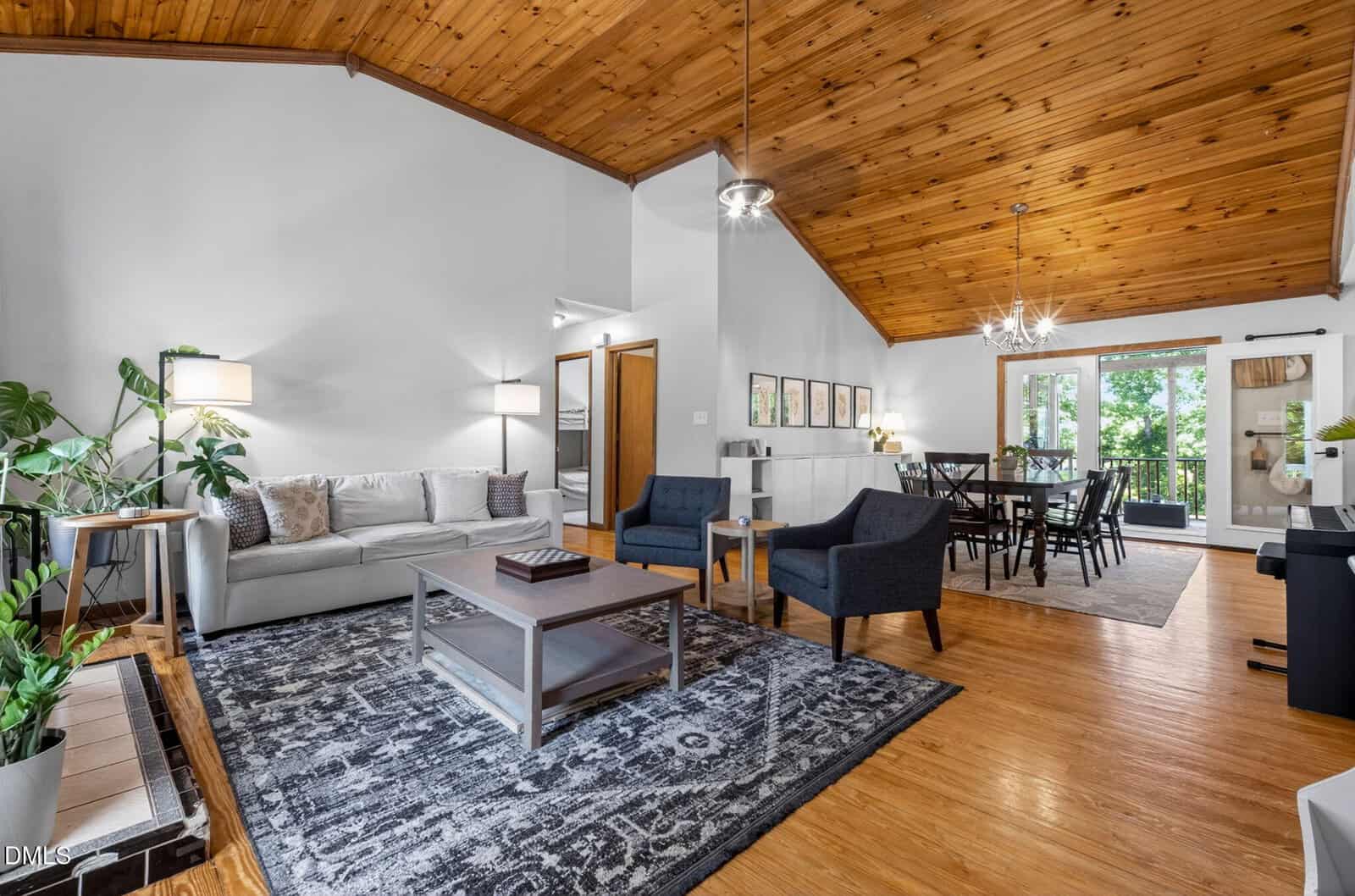 Spacious living and dining area with wood ceiling, white walls, light wood floors, modern furniture, large rug, and glass doors leading to a balcony with greenery outside.