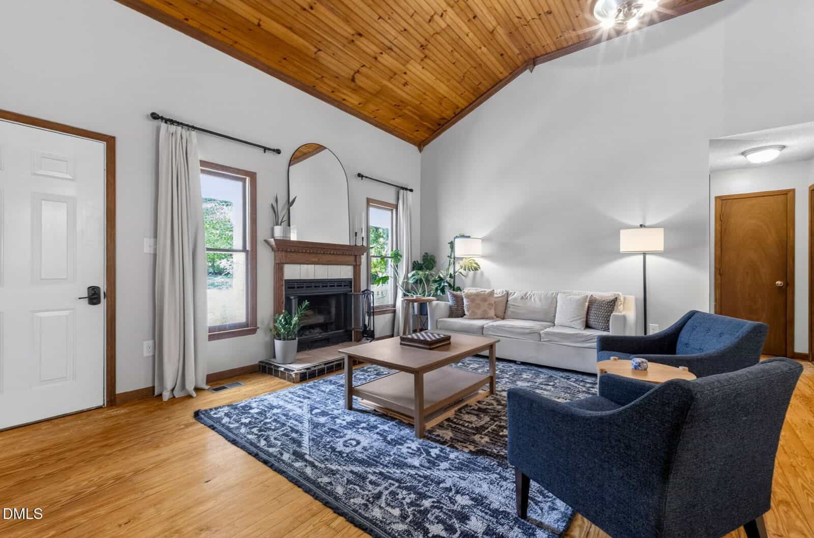 Living room with wood floors, vaulted wood ceiling, and neutral walls. Includes a fireplace, mirror, sofa, two blue chairs, coffee table, rug, tall lamp, and large windows with curtains.