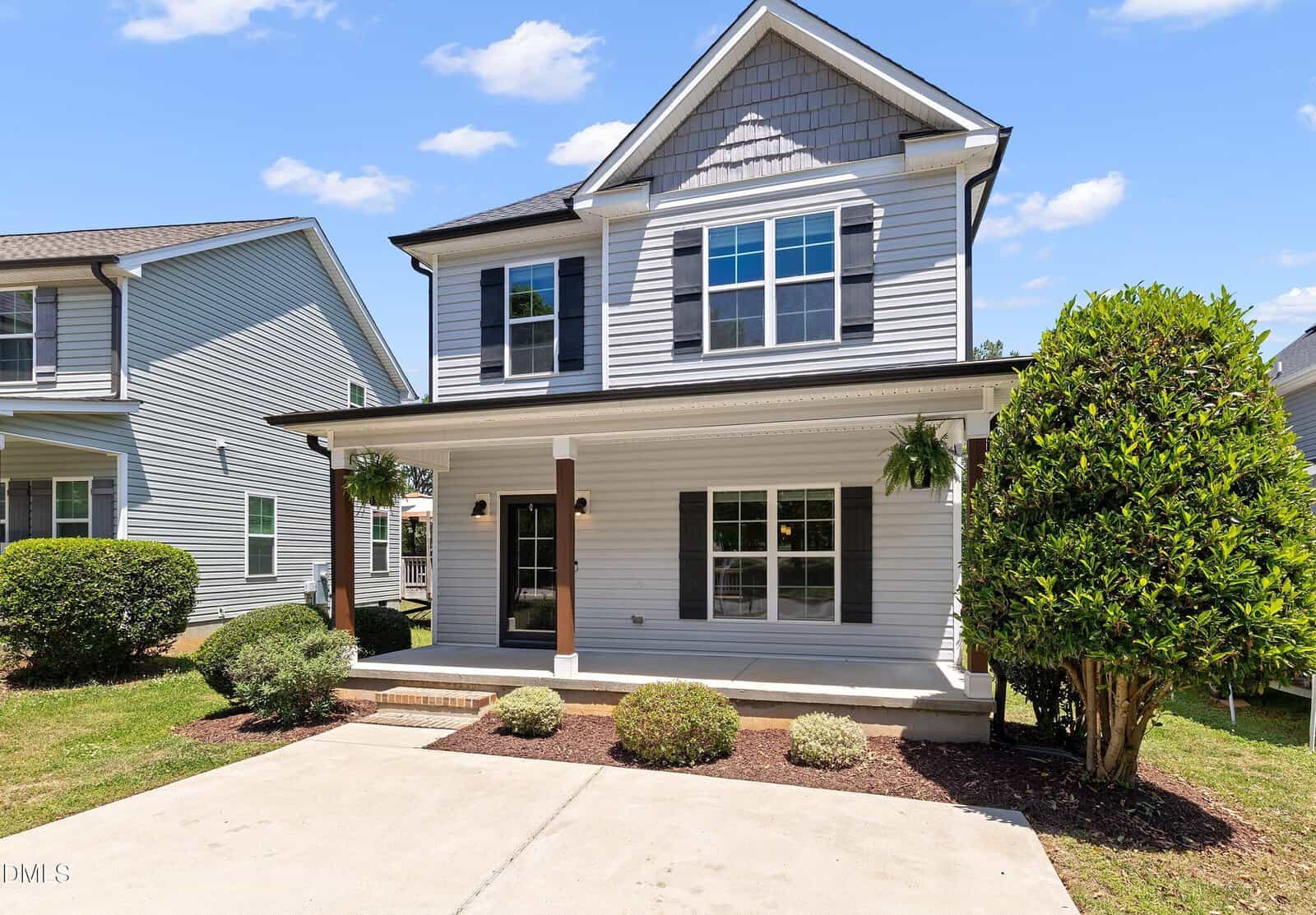 A two-story light gray house with black shutters, a covered front porch, and neatly trimmed bushes and trees. There are hanging plants on the porch and a concrete driveway in front. The sky is clear with a few clouds.