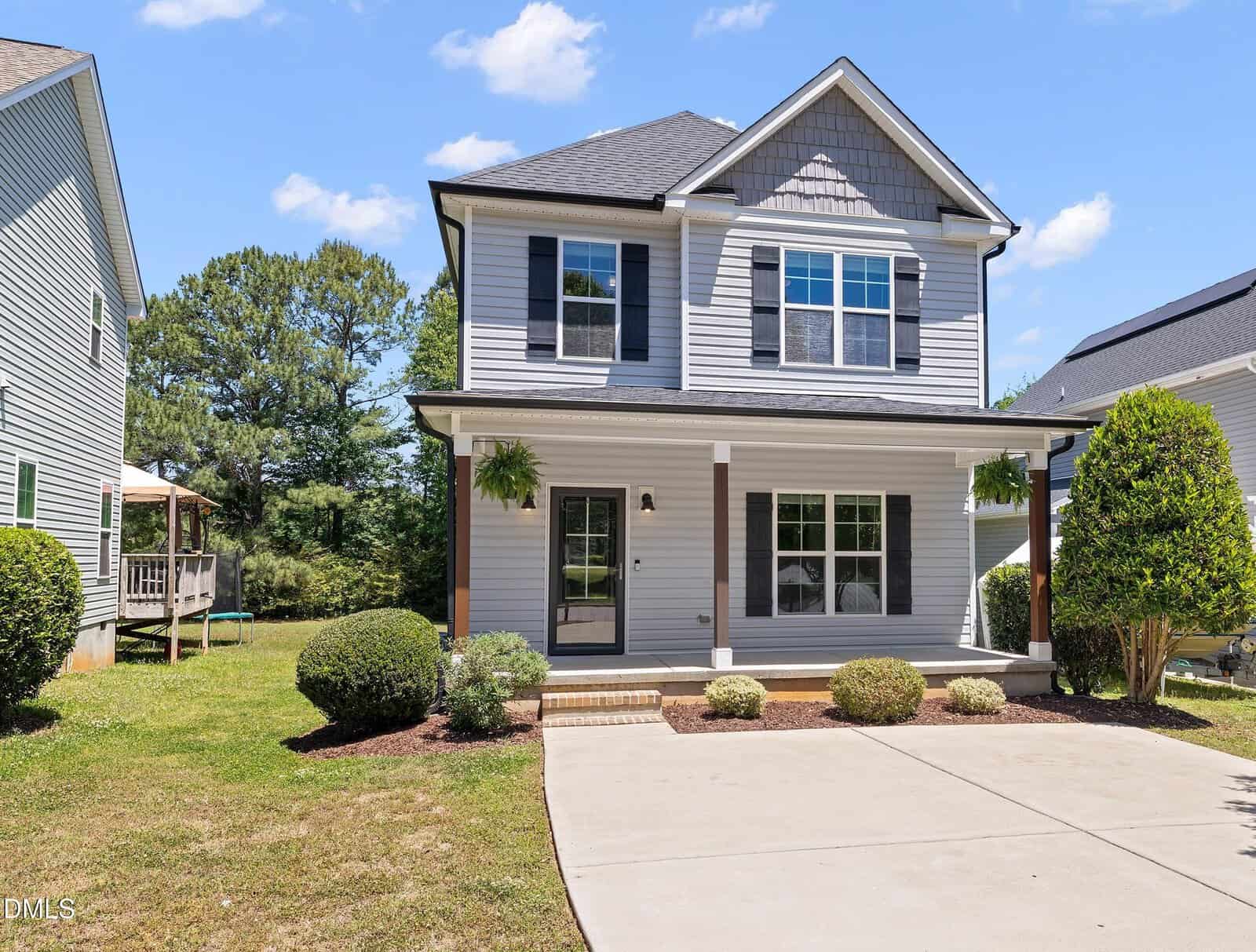 A two-story gray house with white trim, black shutters, a covered front porch, and a driveway. The front yard has neatly trimmed bushes and small trees, with a grassy area and a blue sky overhead.