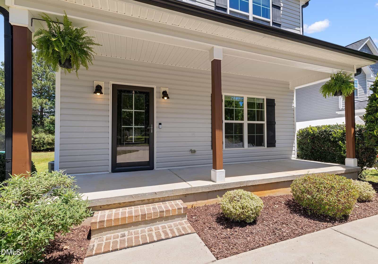 A covered front porch with a black door, two outdoor lights, brown columns, white siding, black shutters, and two hanging ferns. Brick steps and neatly landscaped bushes are in the foreground.