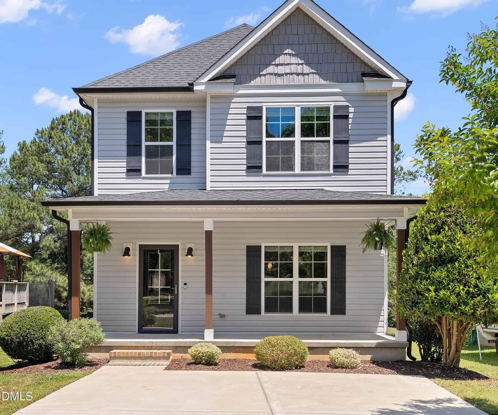 A two-story gray house with white trim at 103 Cottage Court, Youngsville, features a large front porch, black shutters, and a neatly landscaped yard with bushes and hanging plants on a sunny day.