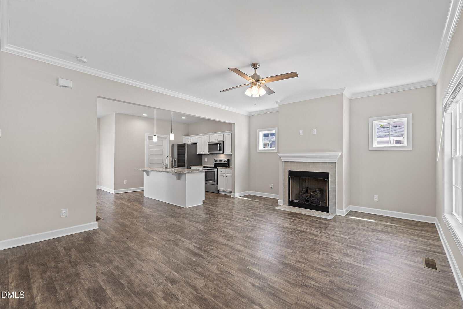 Bright, modern living room with wood floors, a ceiling fan, fireplace, and large windows; open to a white kitchen with an island, stainless steel appliances, and recessed lighting.