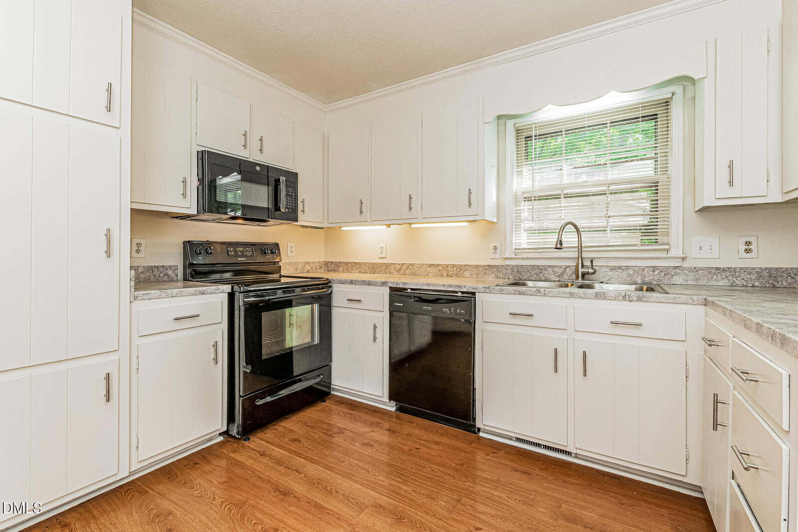 A clean, modern kitchen with white cabinets, black appliances, wood flooring, a double sink under a window with blinds, and light gray countertops.
