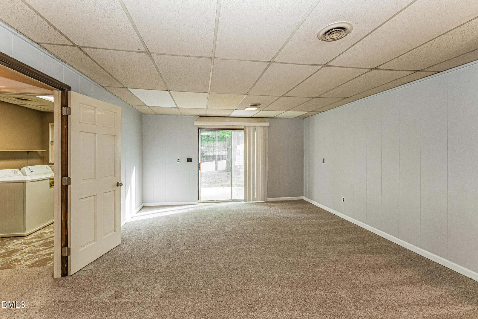 Spacious carpeted basement room with light gray walls, ceiling tiles, sliding glass door leading outside, and open door to a laundry area with washer and dryer.
