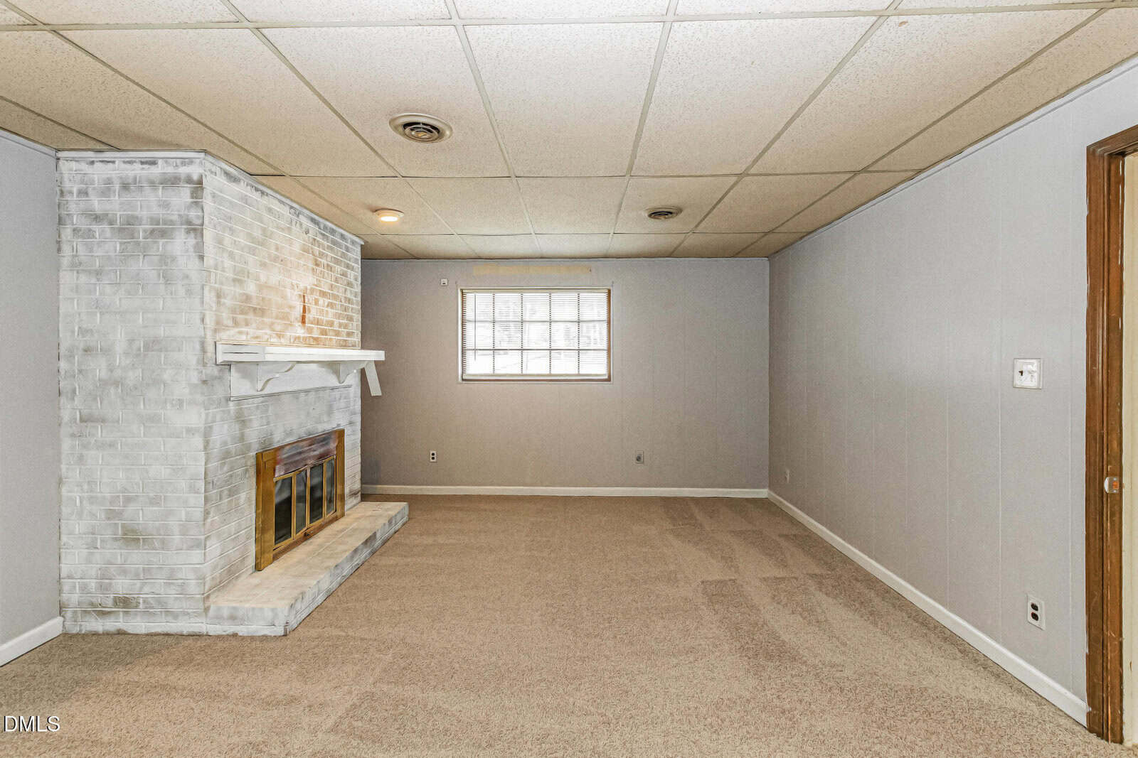 A basement room with beige carpet, light gray walls, a white-painted brick fireplace with a brass cover, and a window letting in natural light. The ceiling is tiled, and the room is mostly empty.