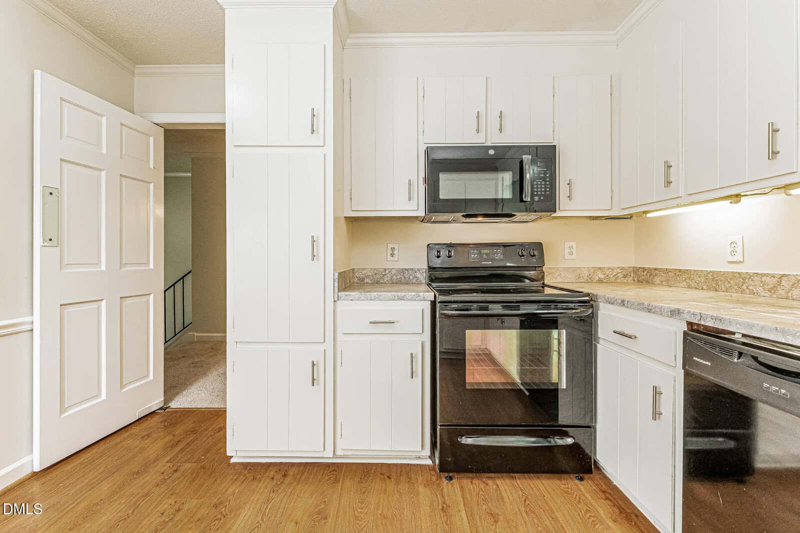 A modern kitchen with white cabinets, black appliances including an oven and microwave, brown speckled countertops, and wood flooring. There's a doorway leading to a carpeted area in the background.