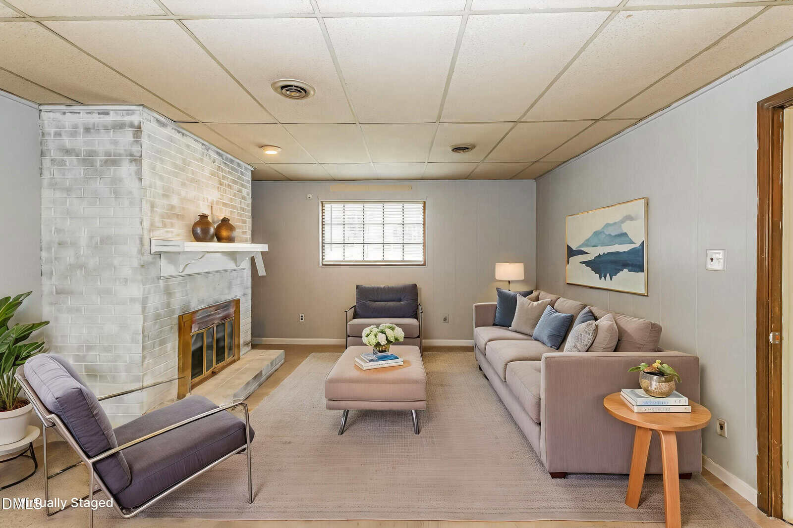 A cozy living room with a gray sofa, armchair, ottoman, and area rug. A white-painted brick fireplace is on the left, with decor on the mantel. A window lets in light, and a landscape picture hangs on the wall.
