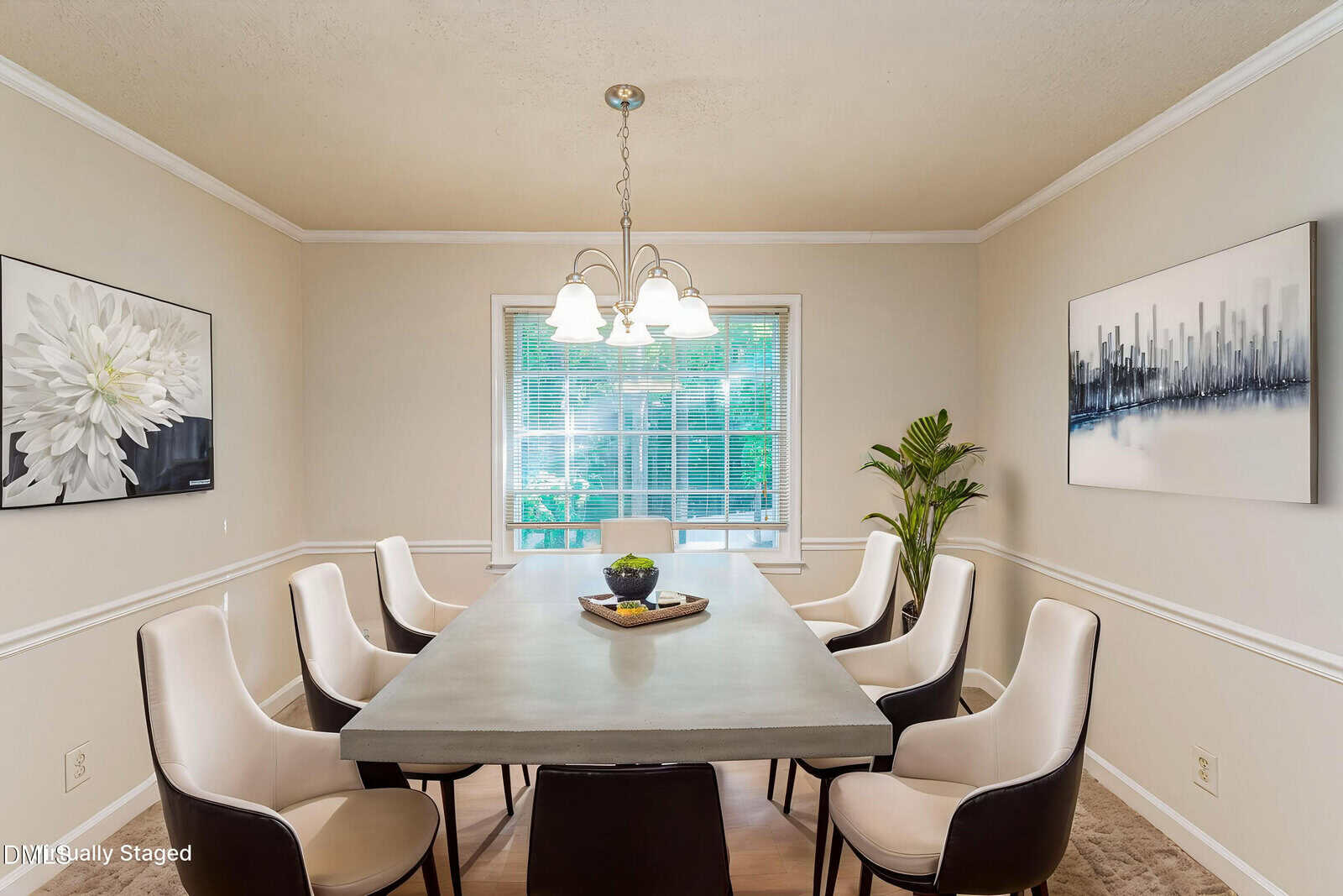 A modern dining room with a rectangular table, six white chairs, a plant in the corner, two large wall art pieces, and a window with blinds letting in natural light. A chandelier hangs above the table.