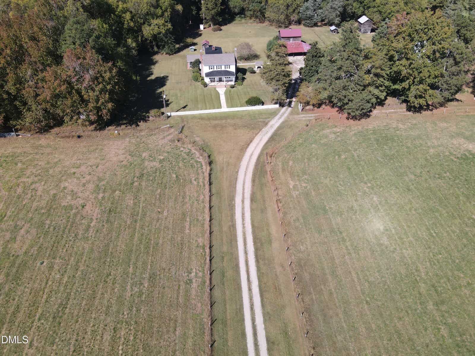 A long gravel driveway leads to a house surrounded by trees and open fields, with a few outbuildings visible near the house, photographed from above on a sunny day.