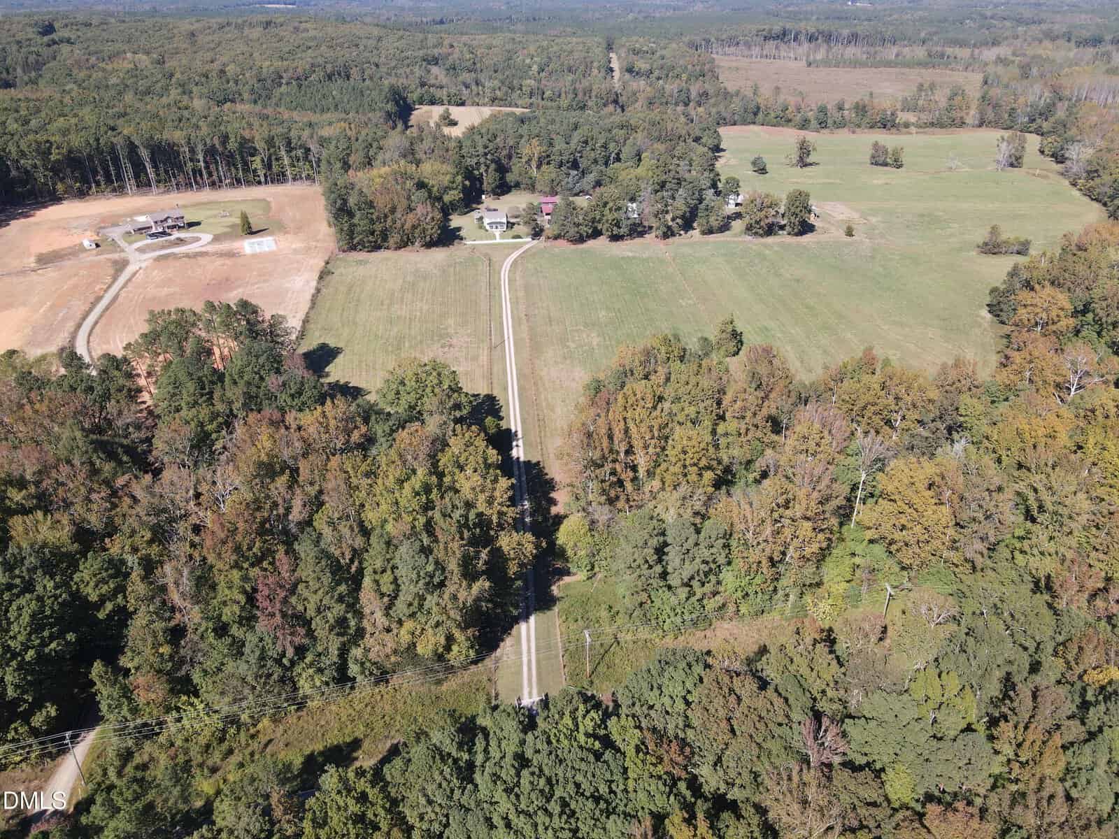 Aerial view of a rural landscape with a straight road running through fields and wooded areas, bordered by scattered houses and farmlands under a clear sky. Dense trees surround open grassy fields.