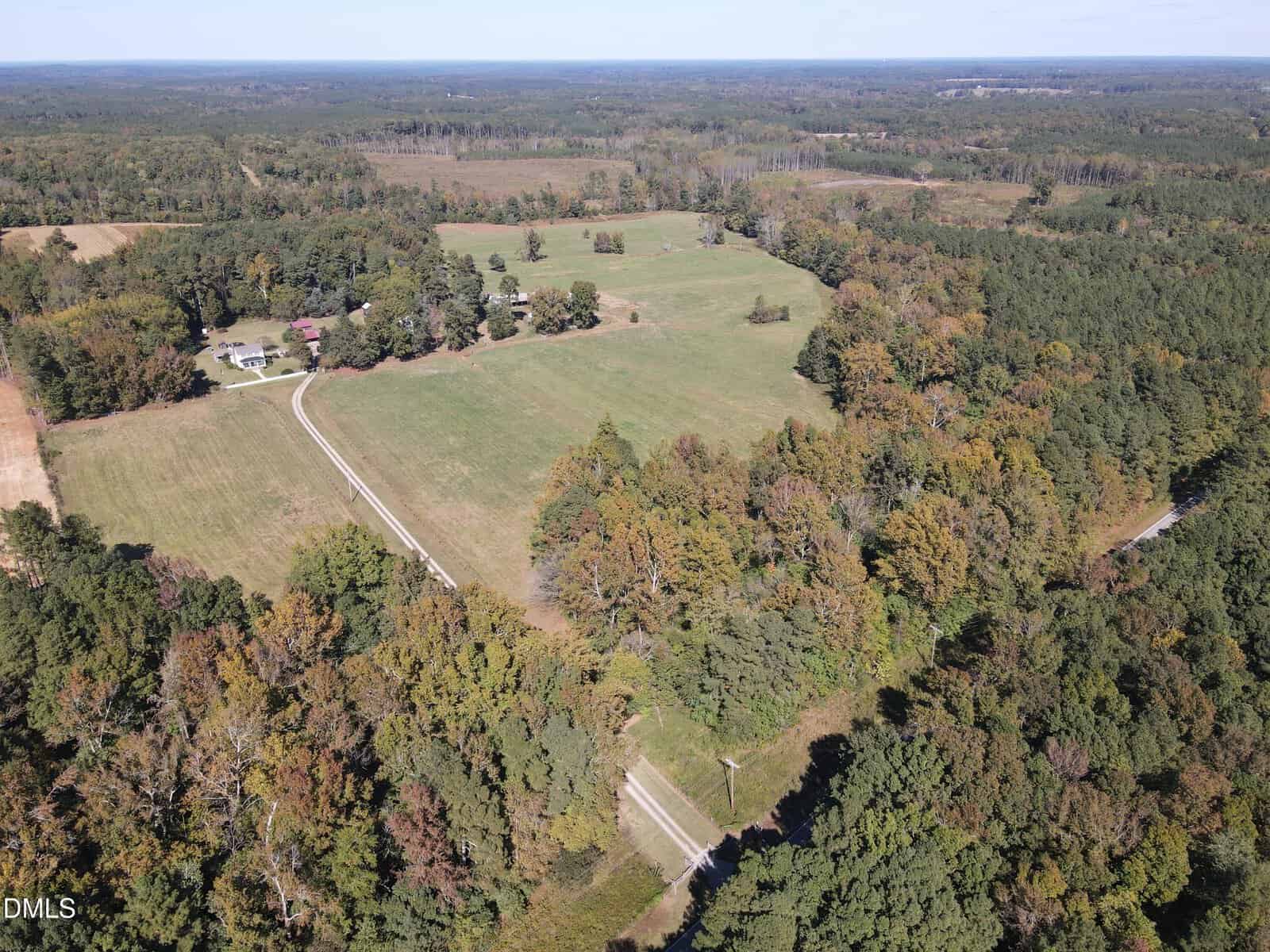Aerial view of a rural landscape featuring open grassy fields bordered by dense forests. A winding road leads to a few houses and farm buildings surrounded by trees under a partly cloudy sky.