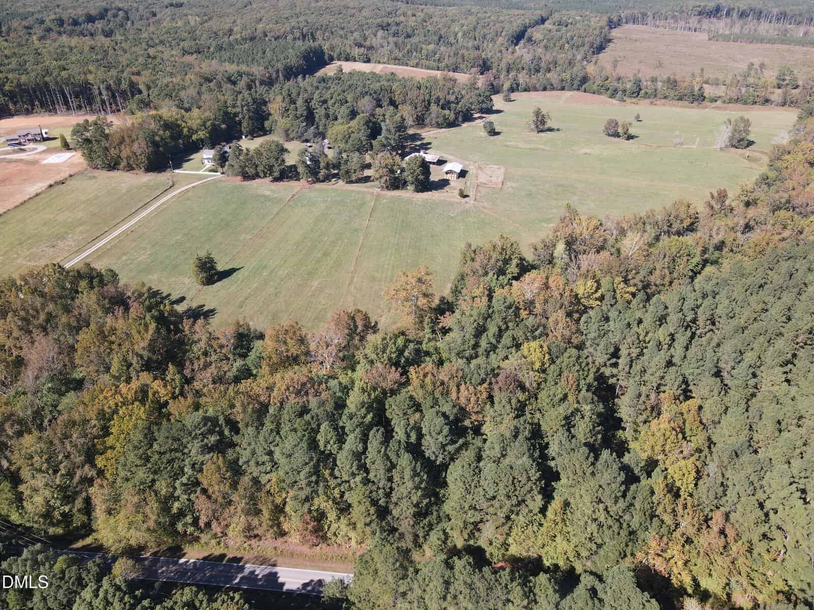 Aerial view of a rural landscape with open fields, scattered trees, a cluster of buildings, and dense forested areas under a clear sky.