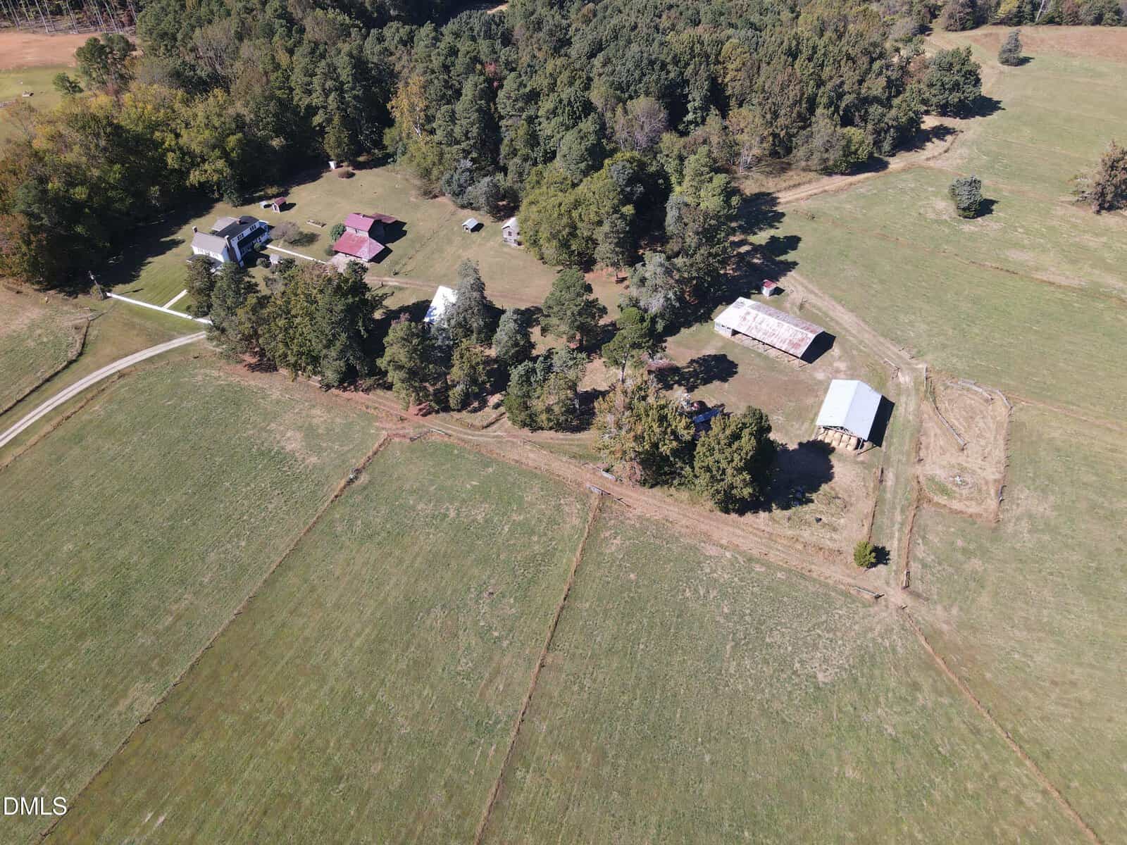 Aerial view of rural property with multiple houses, barns, trees, and open fields bordered by woods, under clear skies.