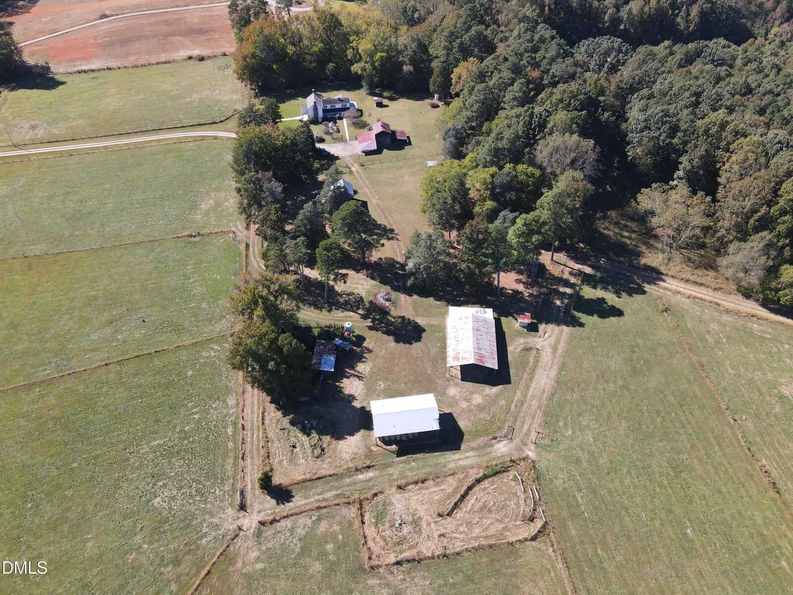 Aerial view of a rural property with two white barns, several smaller buildings, open fields, trees, and a main house surrounded by a driveway. The scene is bordered by wooded areas and farmland.